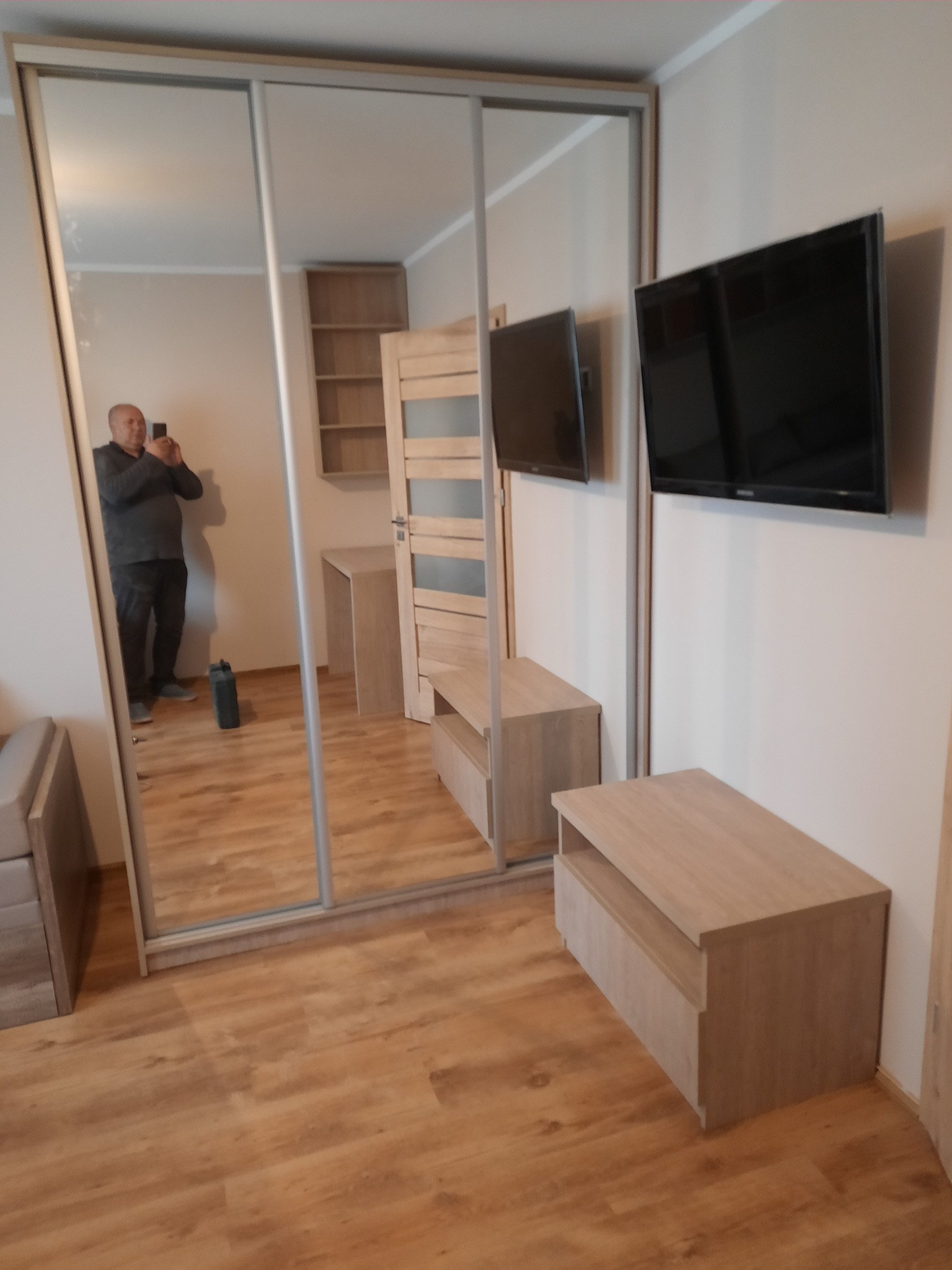 Jasna szafa wnękowa z przesuwnymi lustrzanymi drzwiami, odbijająca wnętrze pokoju z drewnianą podłogą i meblami w odcieniach beżu. Widoczny fragment sofy.