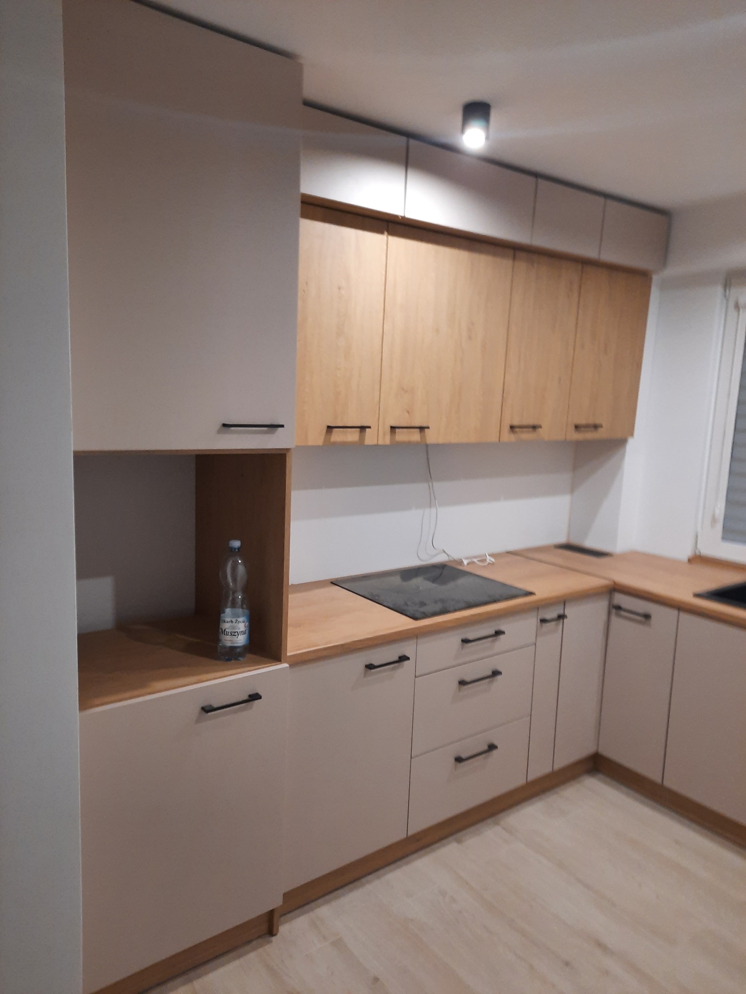 Jasna kuchnia z drewnianymi akcentami: beżowe szafki dolne z czarnymi uchwytami i drewniane szafki górne. Na blacie płyta indukcyjna i butelka wody.