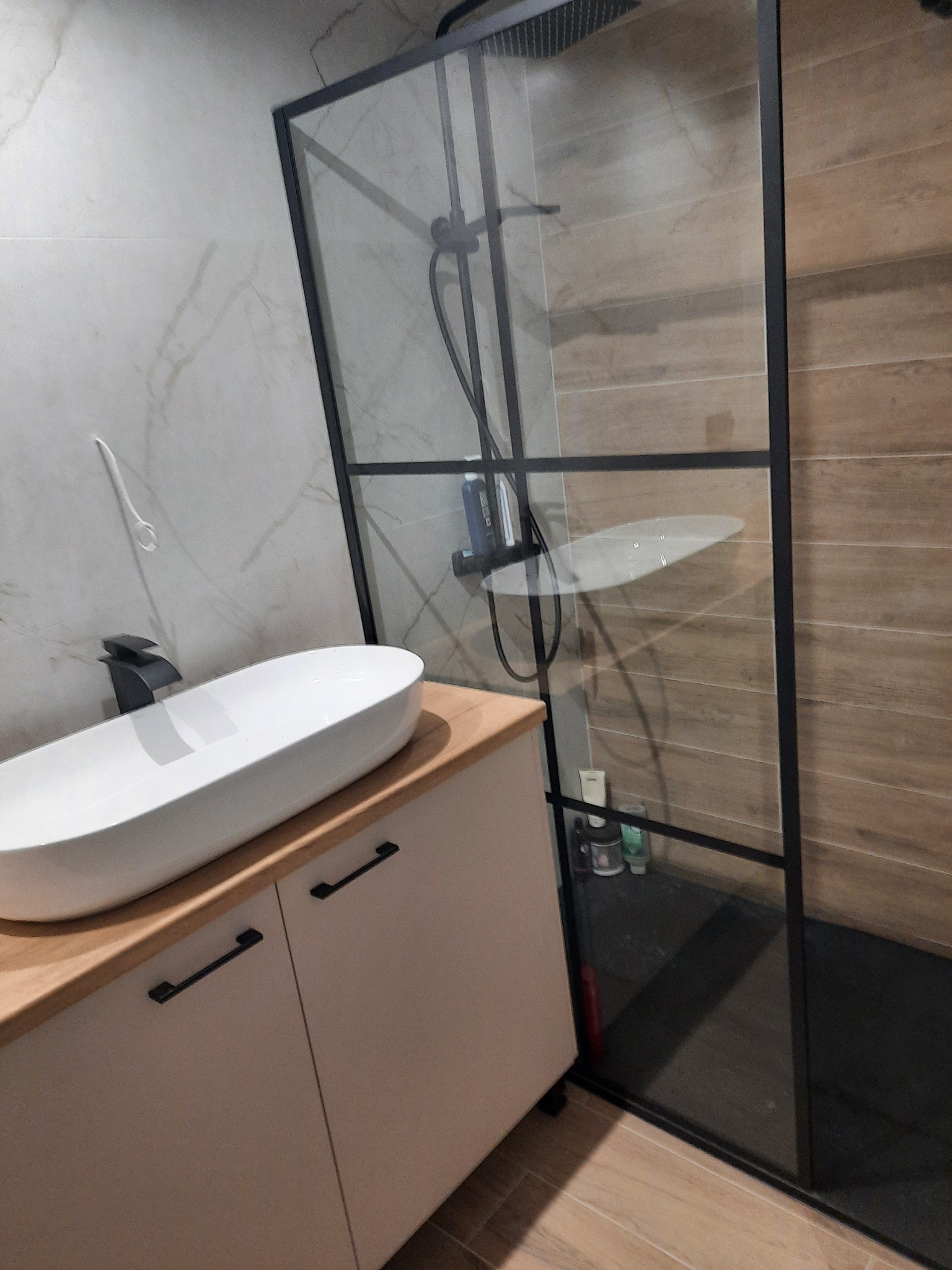 Nowoczesna łazienka z owalną umywalką nablatową na szafce z czarnymi uchwytami i kabiną prysznicową typu walk-in z czarną ramą.