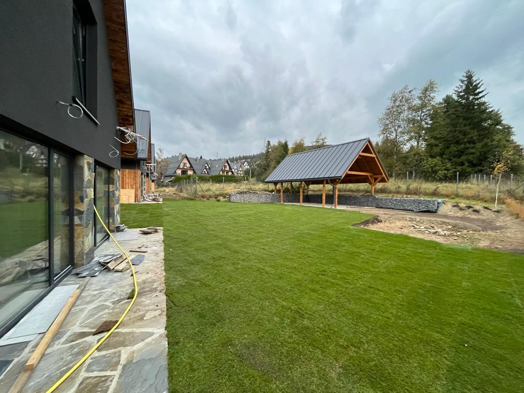 Nowo założony trawnik z rolki przed nowoczesnym domem z kamienną elewacją, w tle drewniana altana i zabudowa z kamienia