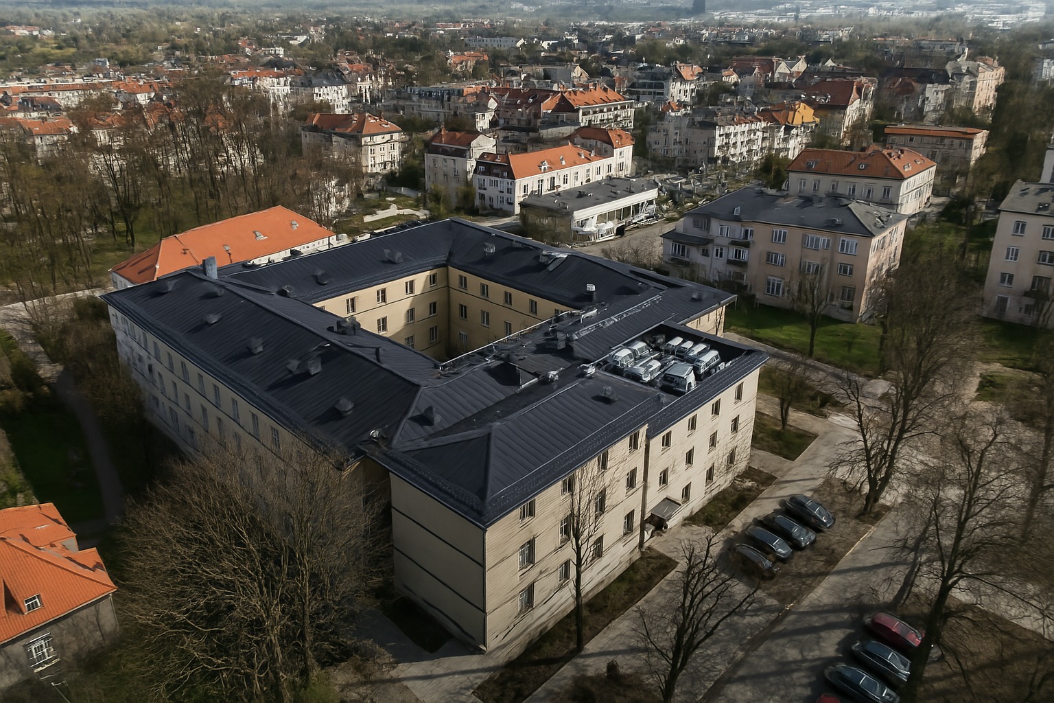Zdjęcie z drona: budynek w kształcie kwadratu z dziedzińcem, ciemny dach, jasna elewacja, zaparkowane samochody na dachu, drzewa bez liści, zabudowa miejska w tle.