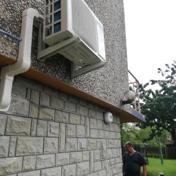 Bartosz Kijas - Biały klimatyzator na elewacji domu z kamienia, widoczne rury odprowadzające. Mężczyzna w ogrodzie, trawa i drzewa w tle. Widok z dołu na urządzenie.