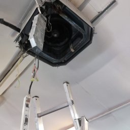 Bartosz Kijas - Otwarta obudowa klimatyzacji kasetonowej na suficie, widoczne okablowanie i drabina, sugerujące prace serwisowe lub instalacyjne.