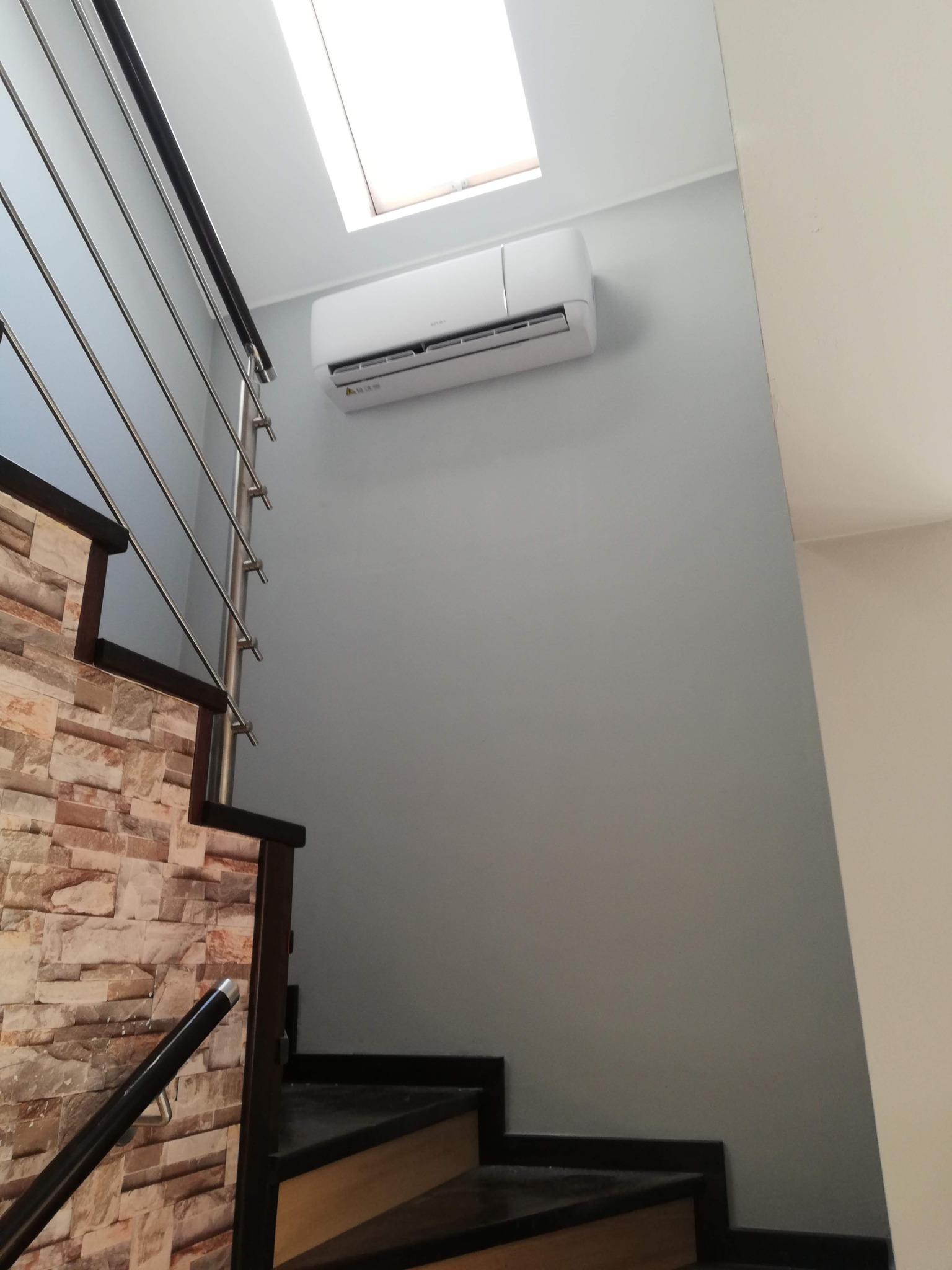 Biały klimatyzator ścienny zamontowany nad schodami z kamienną okładziną w nowoczesnym wnętrzu z oknem dachowym.