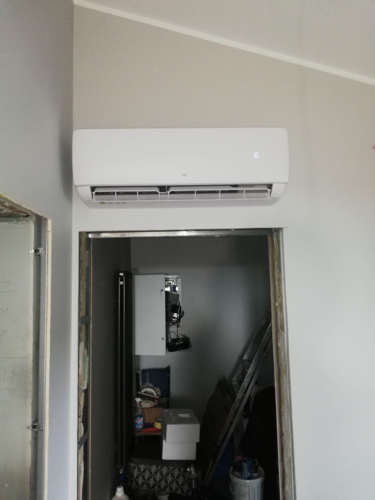 Biała klimatyzacja ścienna zamontowana nad otwartymi drzwiami, widoczne narzędzia i elementy instalacji w pomieszczeniu za drzwiami. Szare ściany.