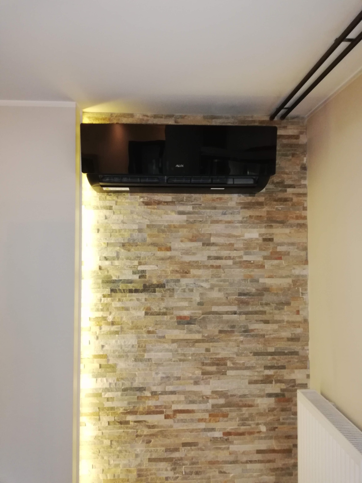 Czarny klimatyzator AUX na ścianie z kamienia dekoracyjnego, z subtelnym oświetleniem LED po lewej stronie. Widoczny fragment grzejnika i rury pod sufitem.
