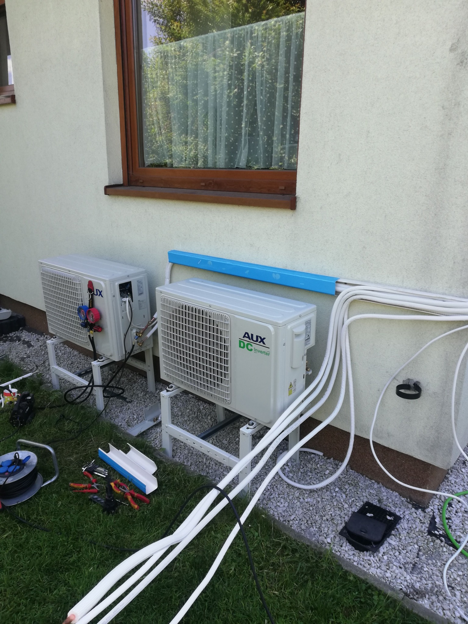 Dwa klimatyzatory AUX DC Inverter zamontowane na zewnątrz budynku, z widocznymi przewodami i narzędziami instalacyjnymi na trawie i żwirze. Widok z dołu.