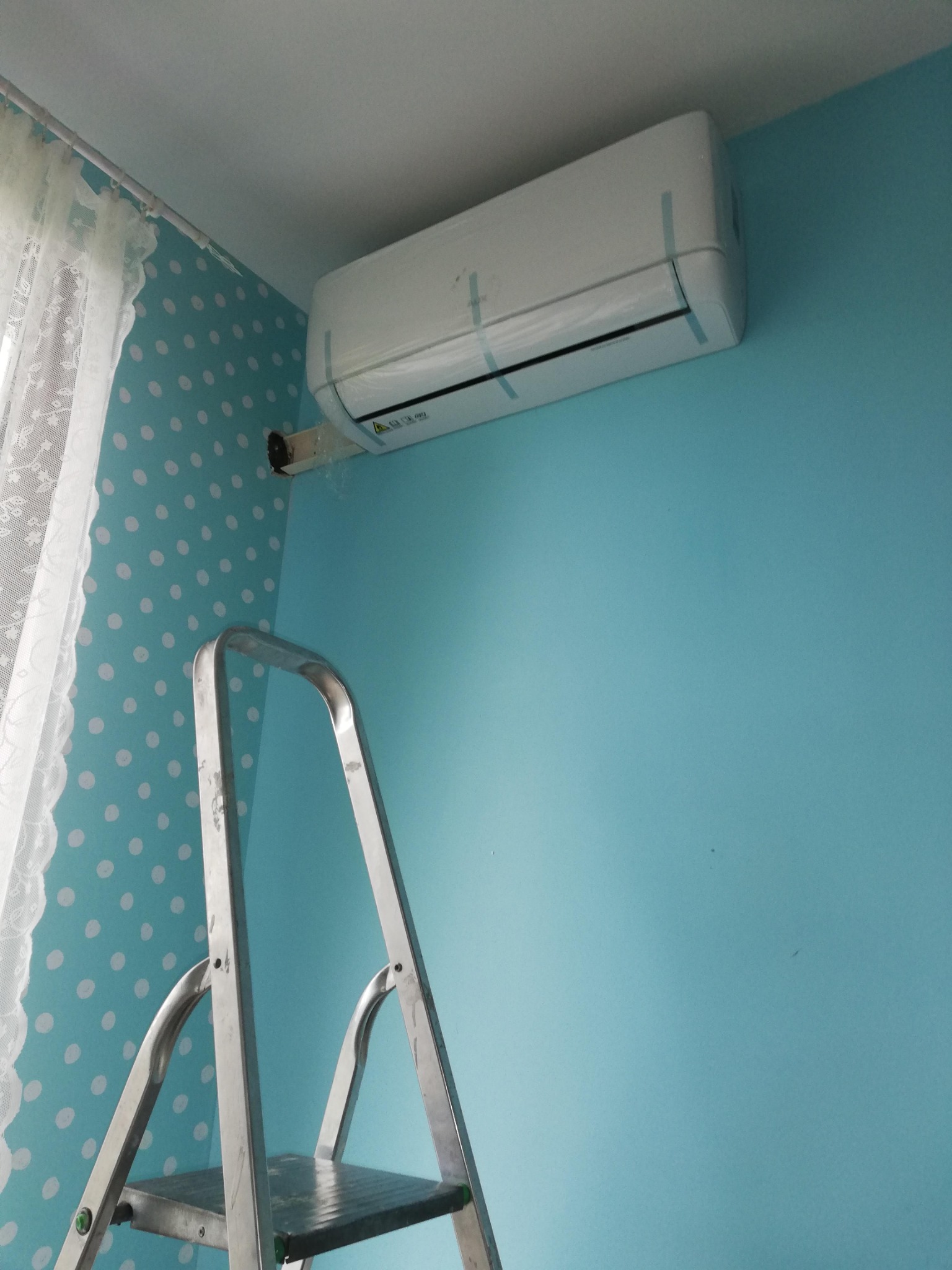 Biała klimatyzacja na błękitnej ścianie, widoczna drabina. Świeżo zamontowane urządzenie zabezpieczone taśmą, na tle ściany w kropki i zasłony.