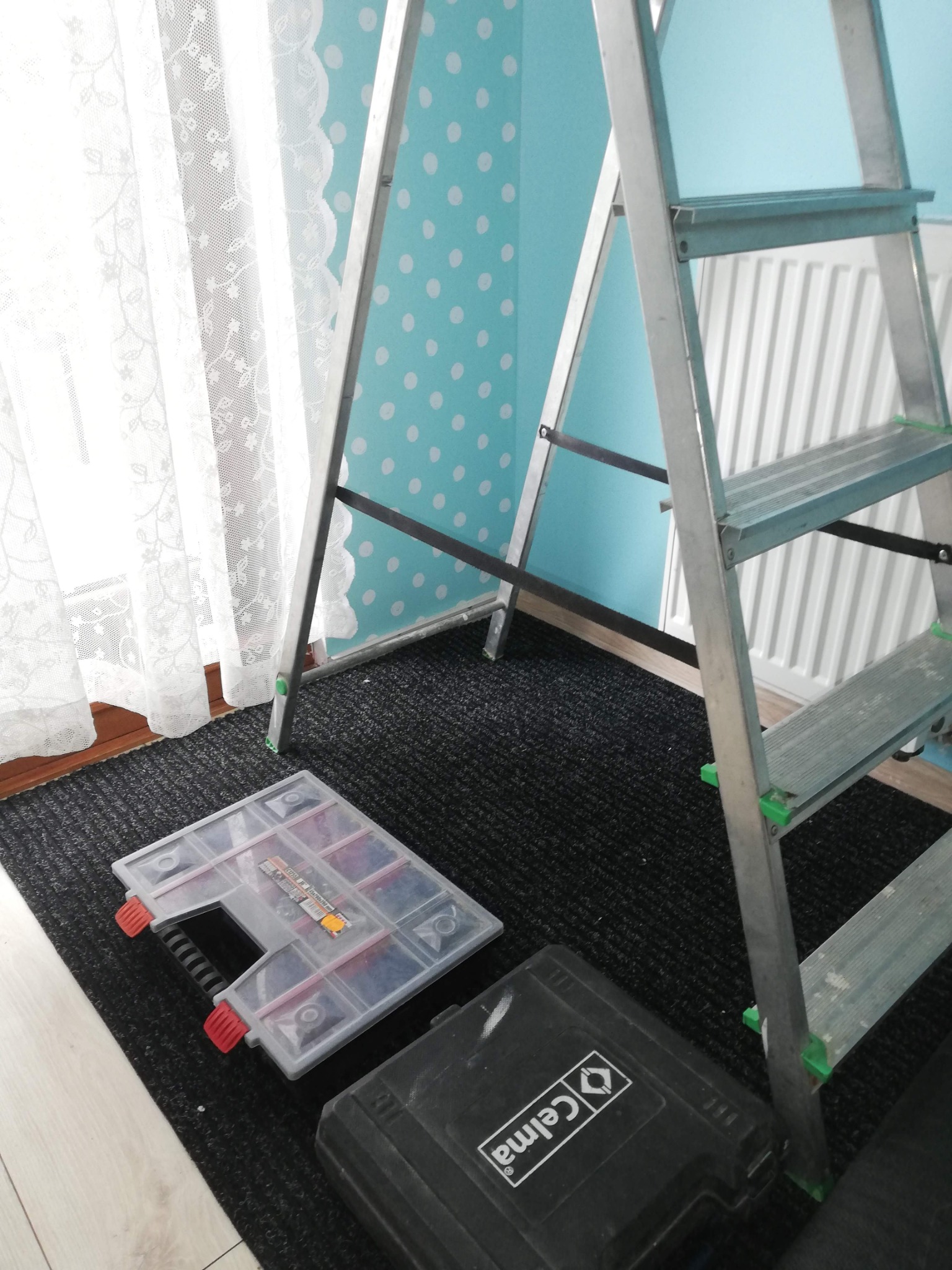 Rozstawiona aluminiowa drabina w pokoju z turkusową ścianą w białe kropki. Na podłodze skrzynki narzędziowe i dywanik. Widok na okno z białymi zasłonami.