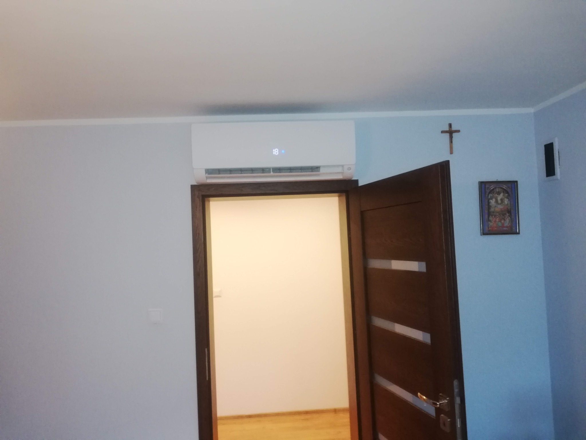 Biały klimatyzator ścienny z wyświetlaczem 18 stopni, zamontowany nad otwartymi drzwiami w pokoju z krzyżem na ścianie.
