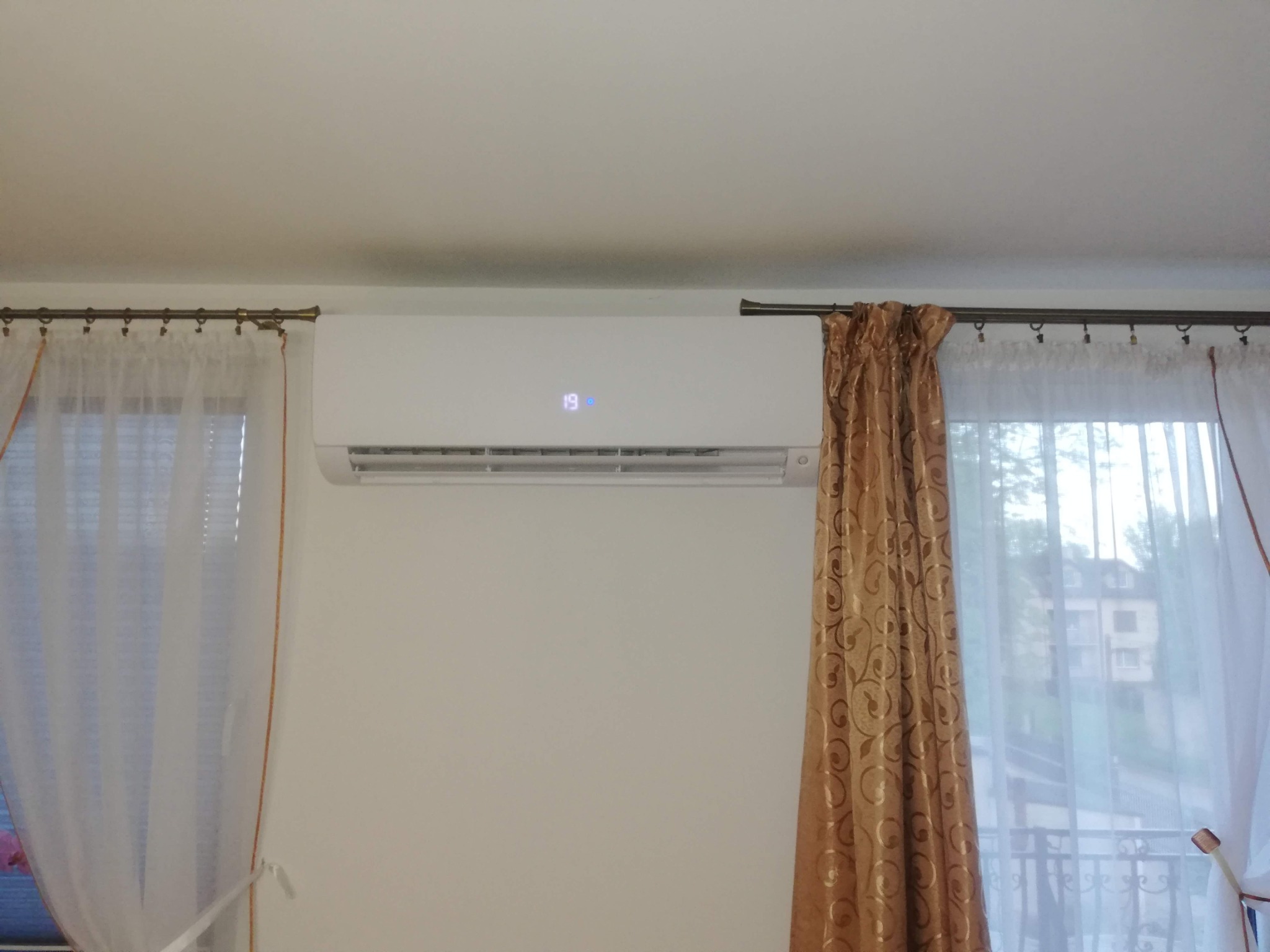 Biały klimatyzator ścienny z wyświetlaczem temperatury 19 stopni, zamontowany w pokoju z zasłonami i widokiem na zewnątrz. Gotowy do użytku.