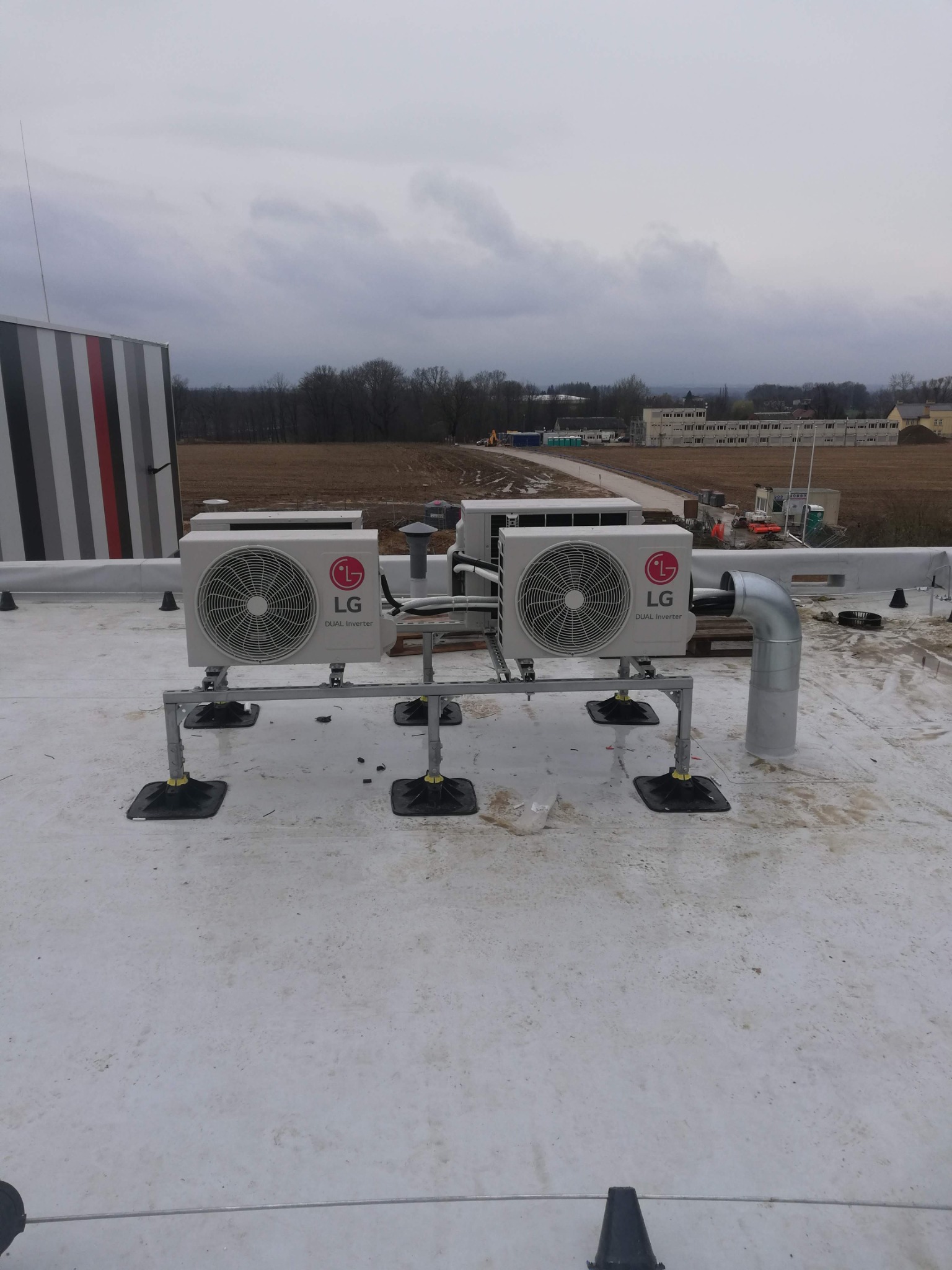 Dwa klimatyzatory LG Dual Inverter na dachu budynku, zamontowane na metalowej konstrukcji. Widoczny system odprowadzania skroplin i elementy mocujące.