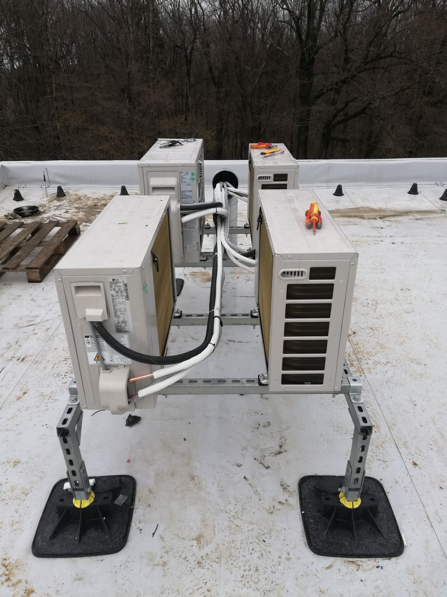 Cztery jednostki zewnętrzne klimatyzacji LG zamontowane na dachu budynku, połączone przewodami, z narzędziami na górze. Widoczne mocowania i elementy instalacji.