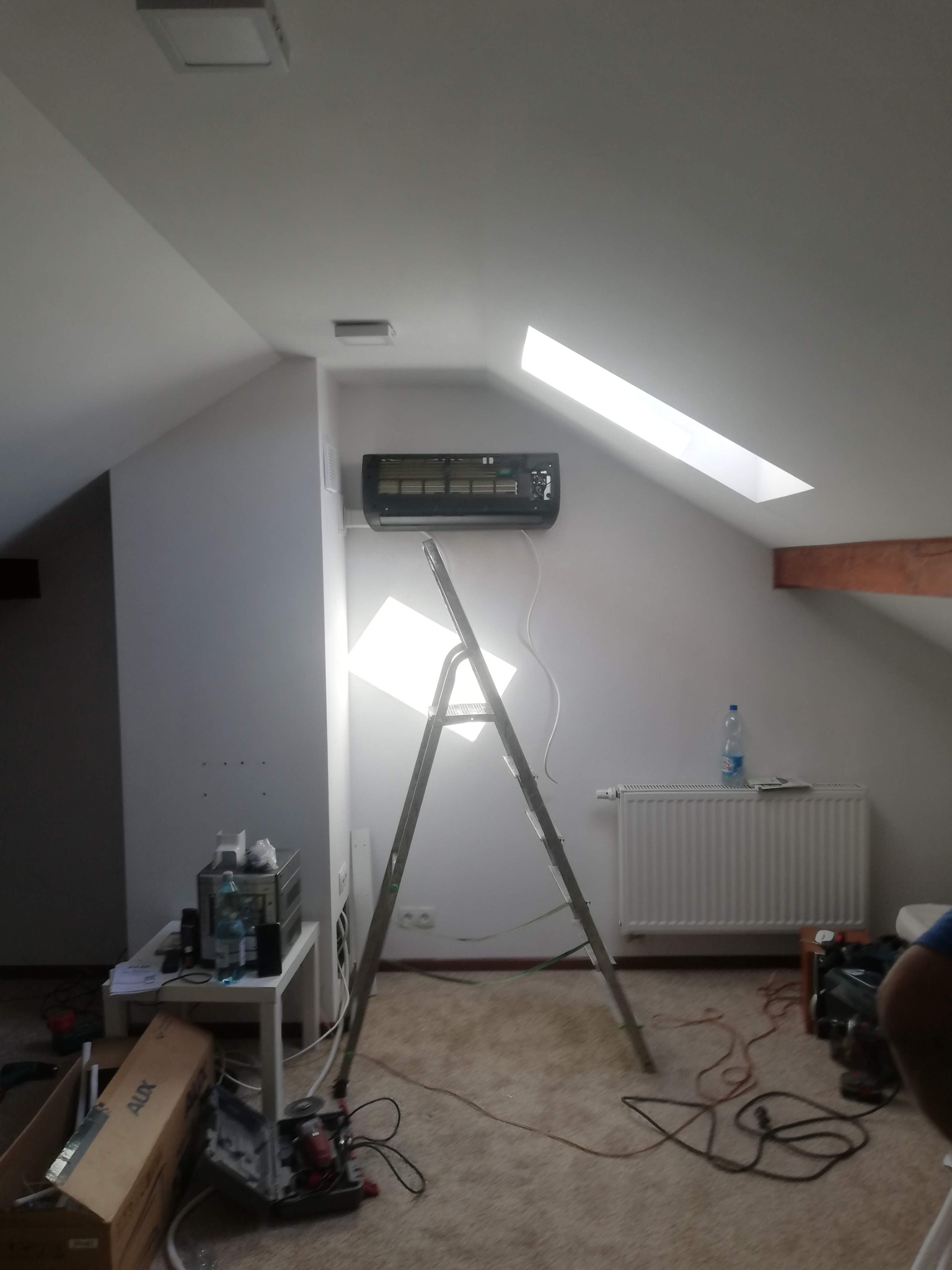 Montaż klimatyzacji w pokoju na poddaszu. Widoczna drabina, jednostka klimatyzacyjna na ścianie, narzędzia i instalacja. W tle okno dachowe i grzejnik.