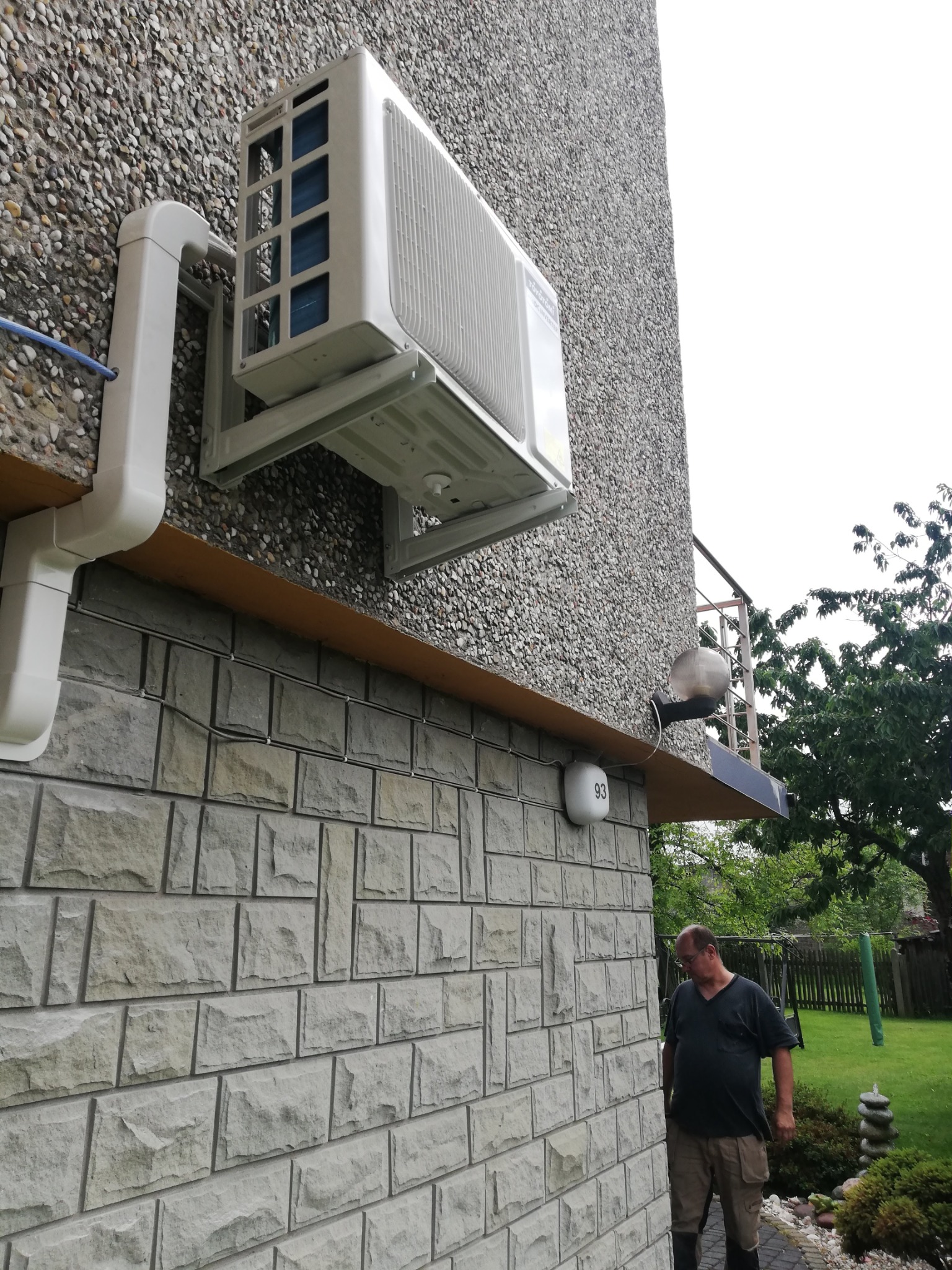 Biały klimatyzator na elewacji domu z kamienia, widoczne rury odprowadzające. Mężczyzna w ogrodzie, trawa i drzewa w tle. Widok z dołu na urządzenie.