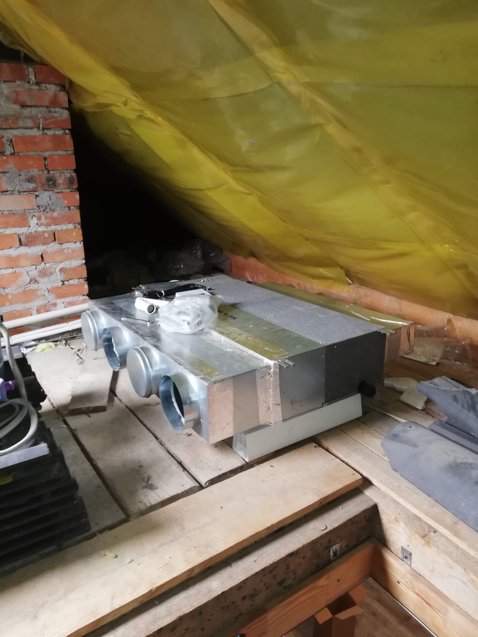 Rekuperator w przestrzeni poddasza, widoczne rury wentylacyjne, ceglana ściana i żółta folia izolacyjna. Instalacja w trakcie montażu.