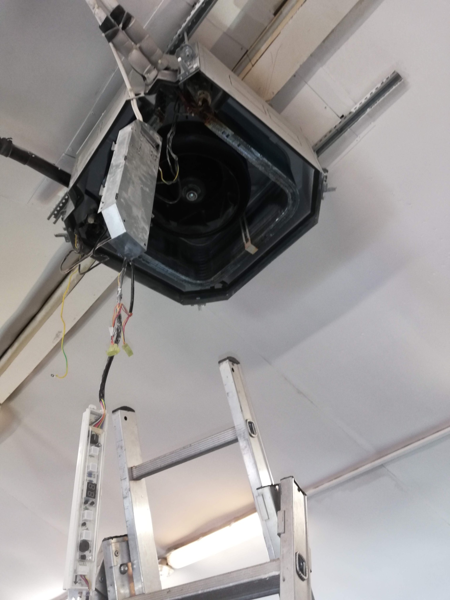 Otwarta obudowa klimatyzacji kasetonowej na suficie, widoczne okablowanie i drabina, sugerujące prace serwisowe lub instalacyjne.