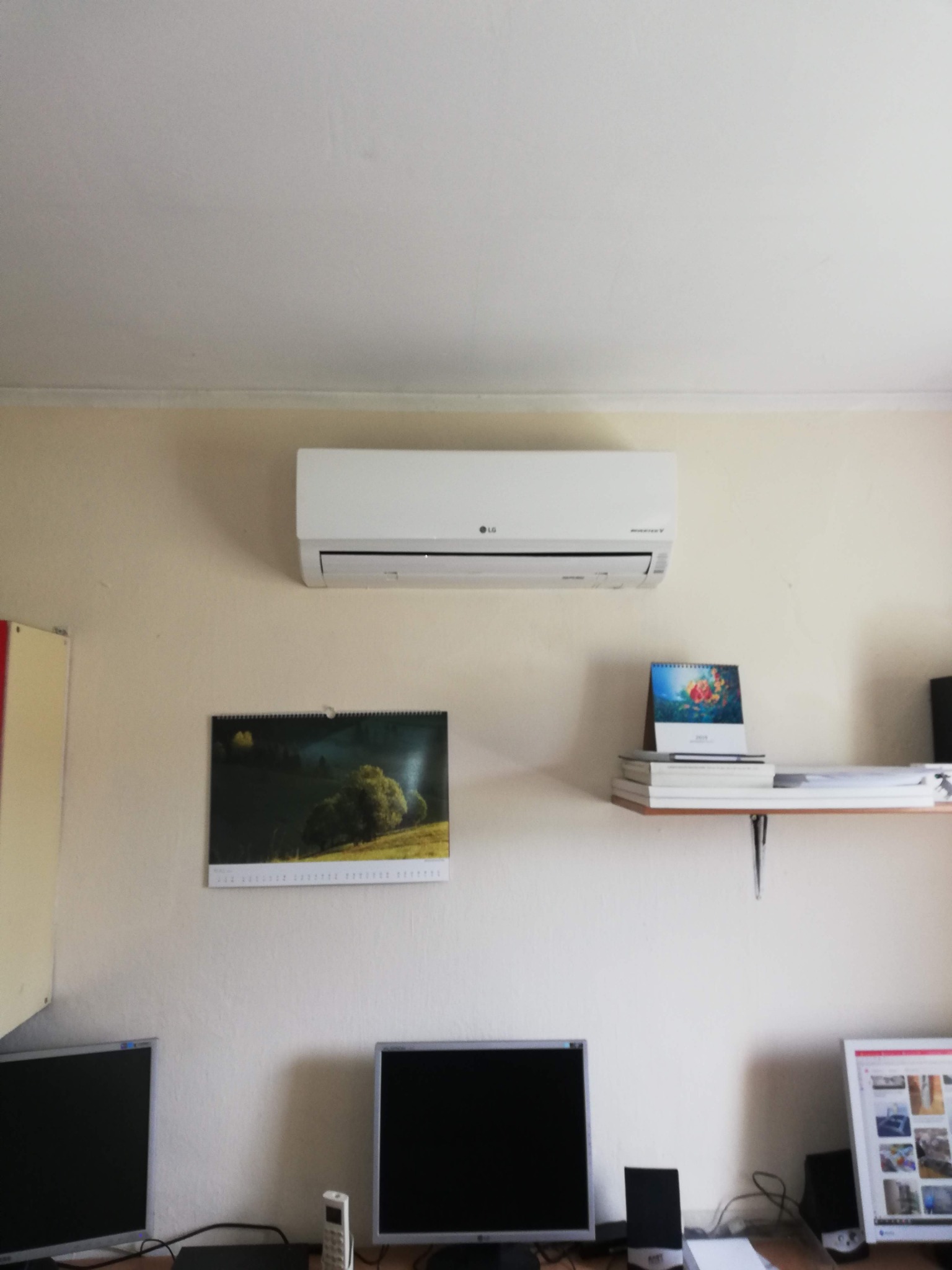 Biała klimatyzacja LG zamontowana na ścianie nad biurkiem z dwoma monitorami i kalendarzami. Widok z przodu, w pomieszczeniu biurowym.