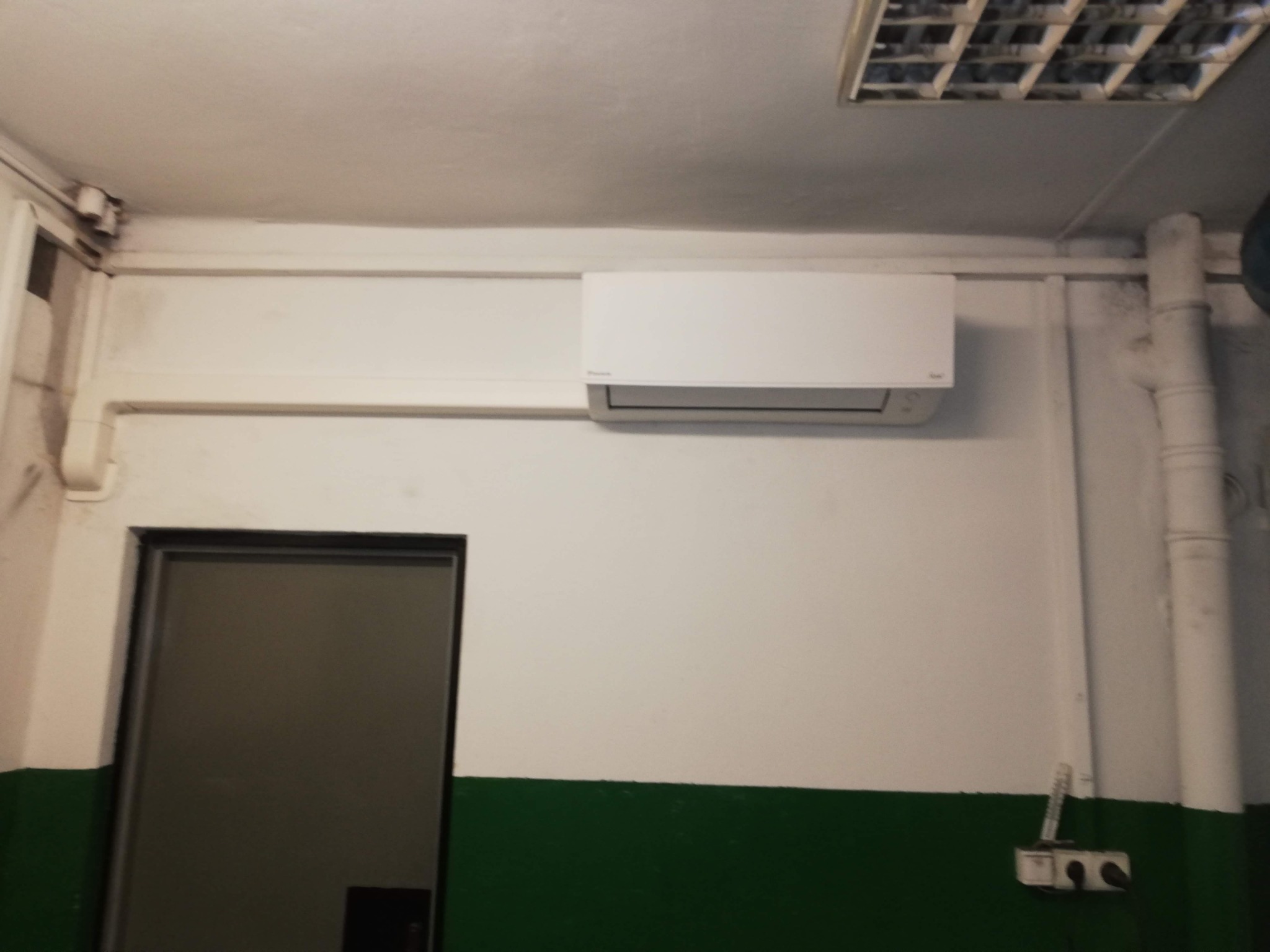 Biały klimatyzator Daikin zamontowany na ścianie nad drzwiami, widoczne rury instalacyjne i biało-zielona lamperia w pomieszczeniu.