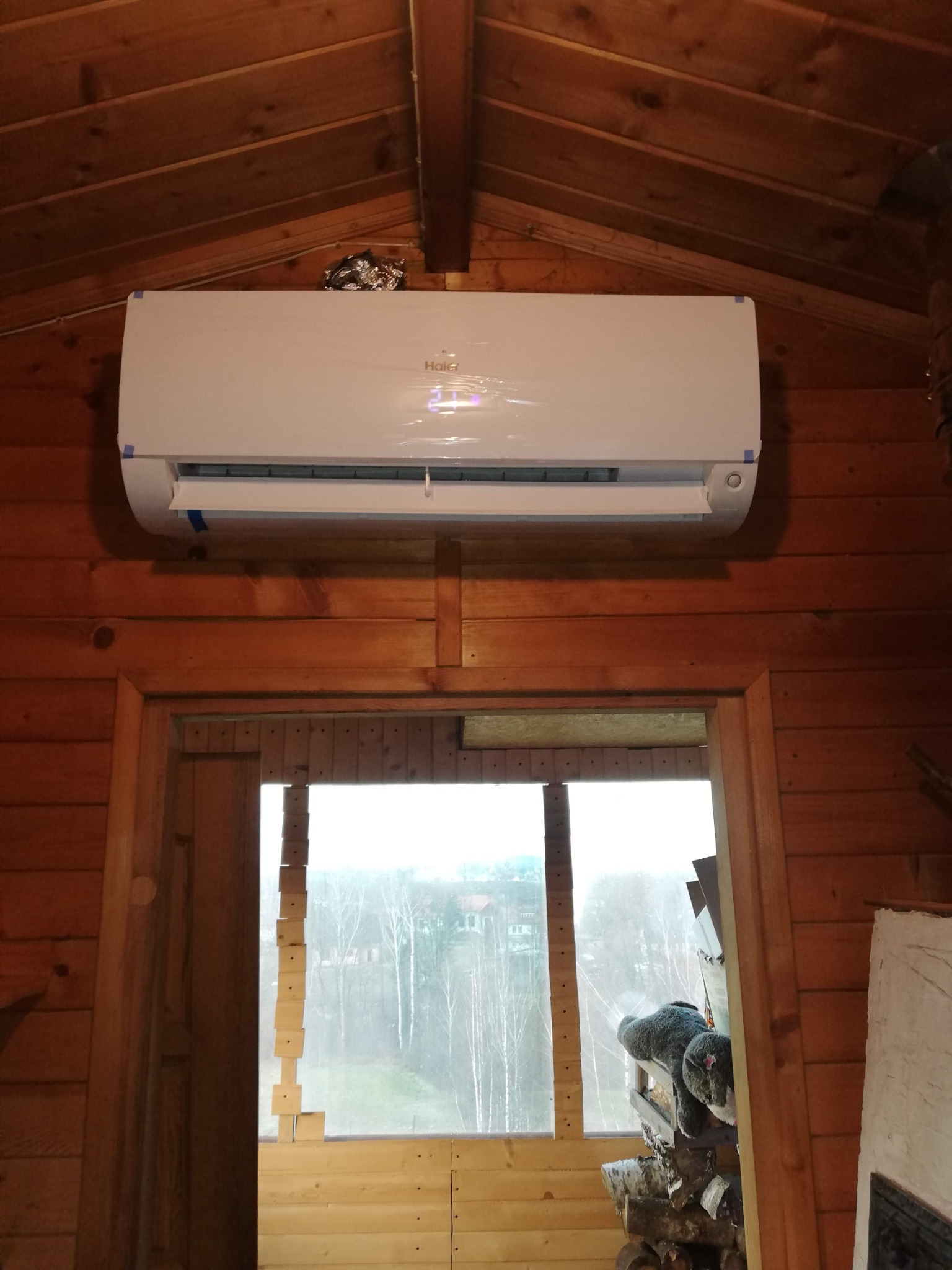 Biały klimatyzator Haier zamontowany na drewnianej ścianie w domku, widok z dołu. Przez okno widoczne drzewa i budynki w oddali.
