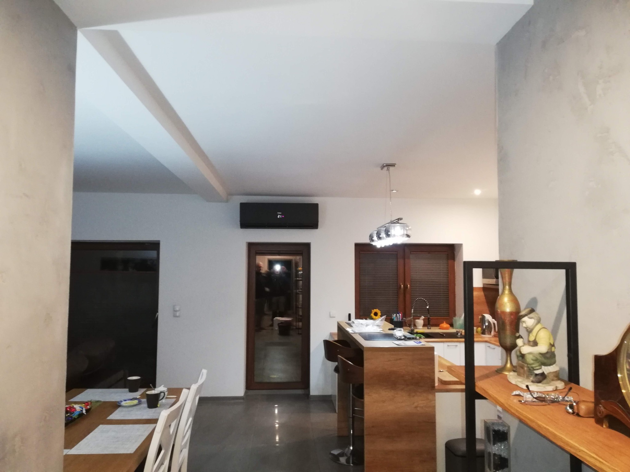 Nowoczesny salon z czarną klimatyzacją ścienną, widok na otwartą kuchnię i jadalnię. Wnętrze w neutralnej kolorystyce z akcentami drewna. Oświetlenie sufitowe.