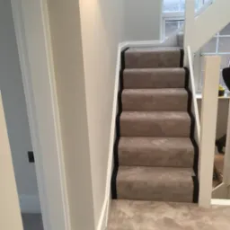 Jasnoszara wykładzina dywanowa na schodach z czarnym obramowaniem, białe ściany i balustrada, widok z dołu schodów.