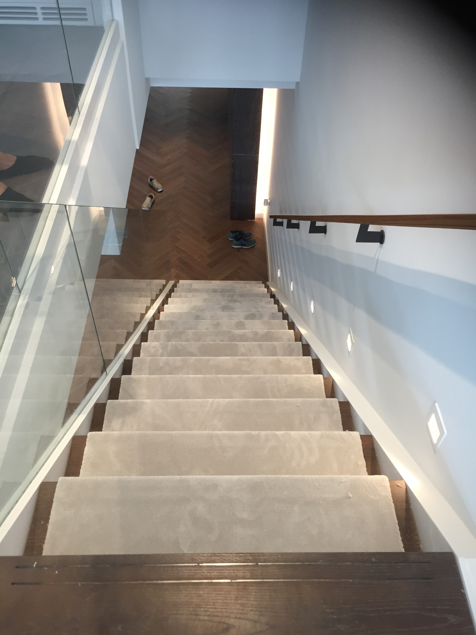 Schody z drewnianymi stopniami pokryte kremową wykładziną dywanową, widok z góry, szklana balustrada, parkiet w jodełkę w tle, buty na podłodze.