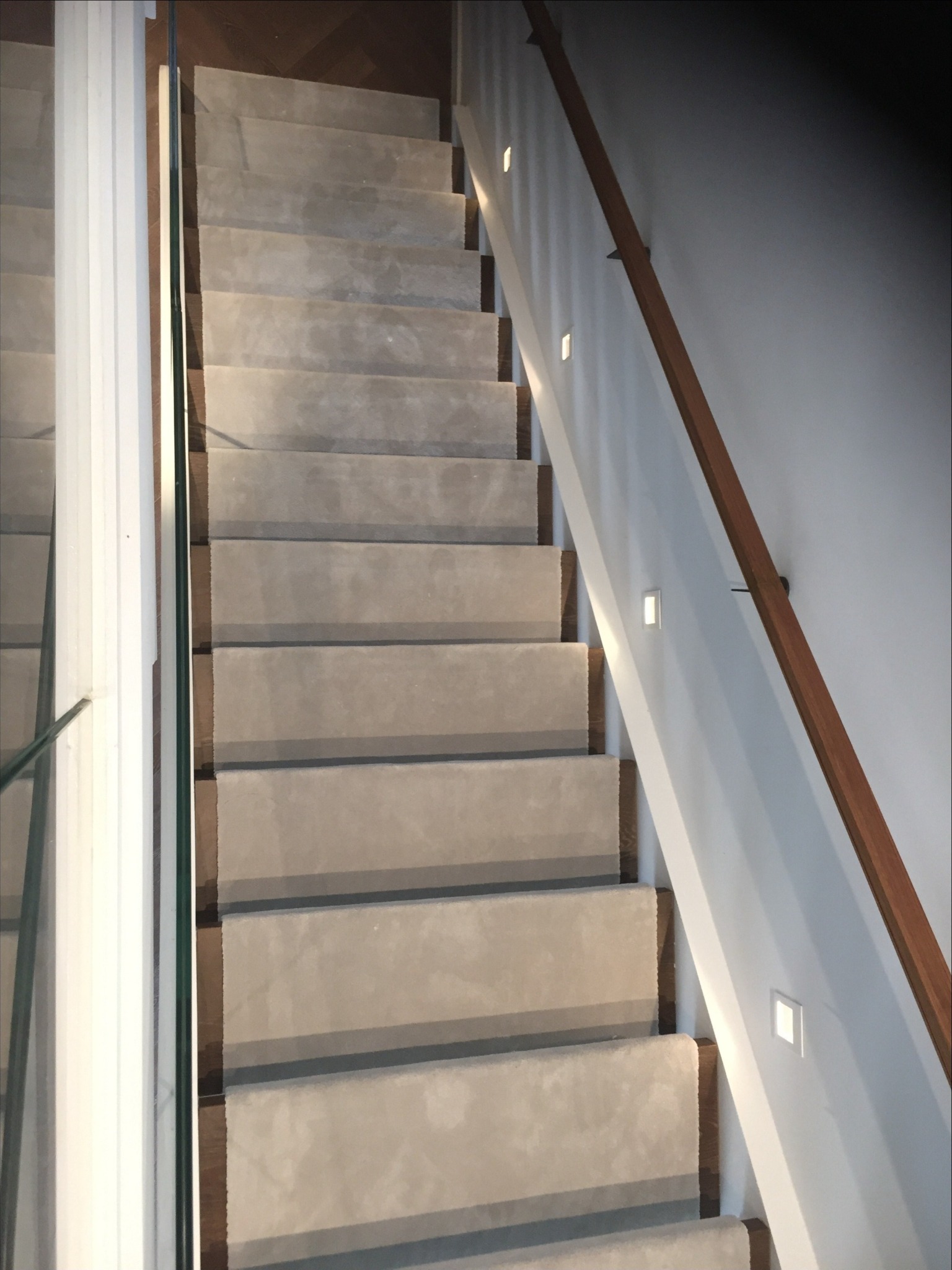 Schody obłożone jasną wykładziną dywanową z drewnianymi krawędziami, oświetlone punktowymi lampami w ścianie i zabezpieczone szklaną balustradą.