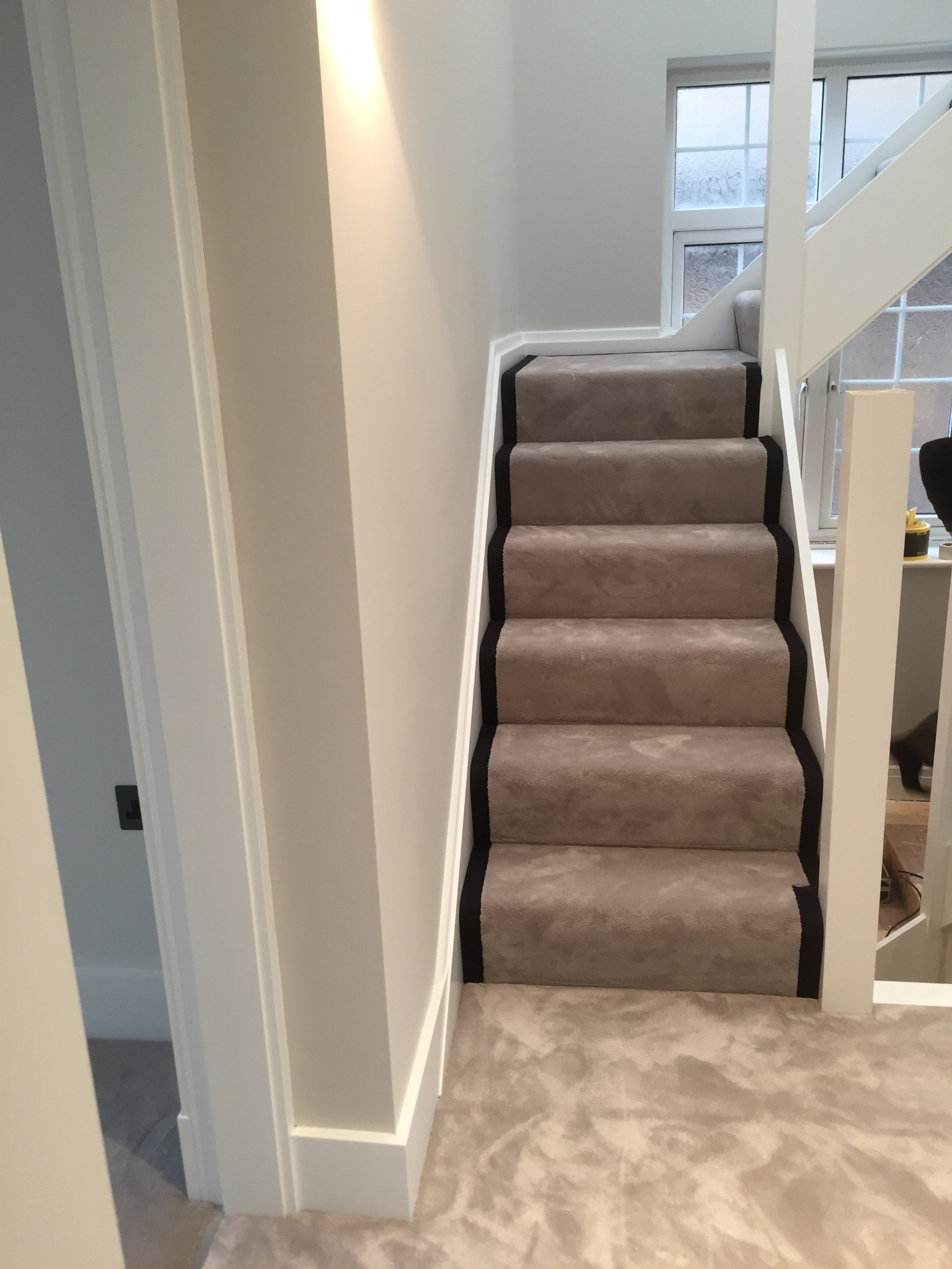 Jasnoszara wykładzina dywanowa na schodach z czarnym obramowaniem, białe ściany i balustrada, widok z dołu schodów.