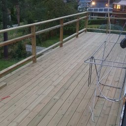 EMKO_BUD EMIL WASILEWSKI - Nowy, drewniany taras z balustradą, widok z góry. W tle zieleń i zabudowania. Na tarasie suszarka na pranie i wystający kabel. Świeże drewno, jasne oświetlenie.