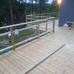 EMKO_BUD EMIL WASILEWSKI - Drewniany taras w trakcie budowy, widoczne deski, balustrady w surowym stanie, narzędzia i materiały budowlane w tle. Budowa tarasu na zewnątrz budynku.