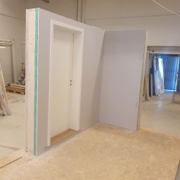 EMKO_BUD EMIL WASILEWSKI - Wnętrze w trakcie prac: fragment ściany z drzwiami i płytą karton gips, podłoga z płyt OSB. Widoczne elementy konstrukcyjne i wykończeniowe. Prace wykończeniowe w toku.