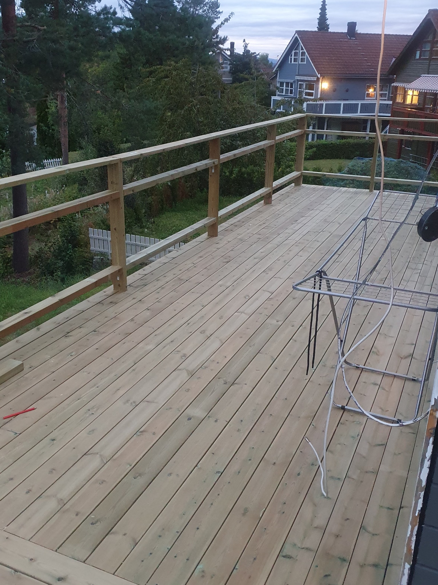 Nowy, drewniany taras z balustradą, widok z góry. W tle zieleń i zabudowania. Na tarasie suszarka na pranie i wystający kabel. Świeże drewno, jasne oświetlenie.