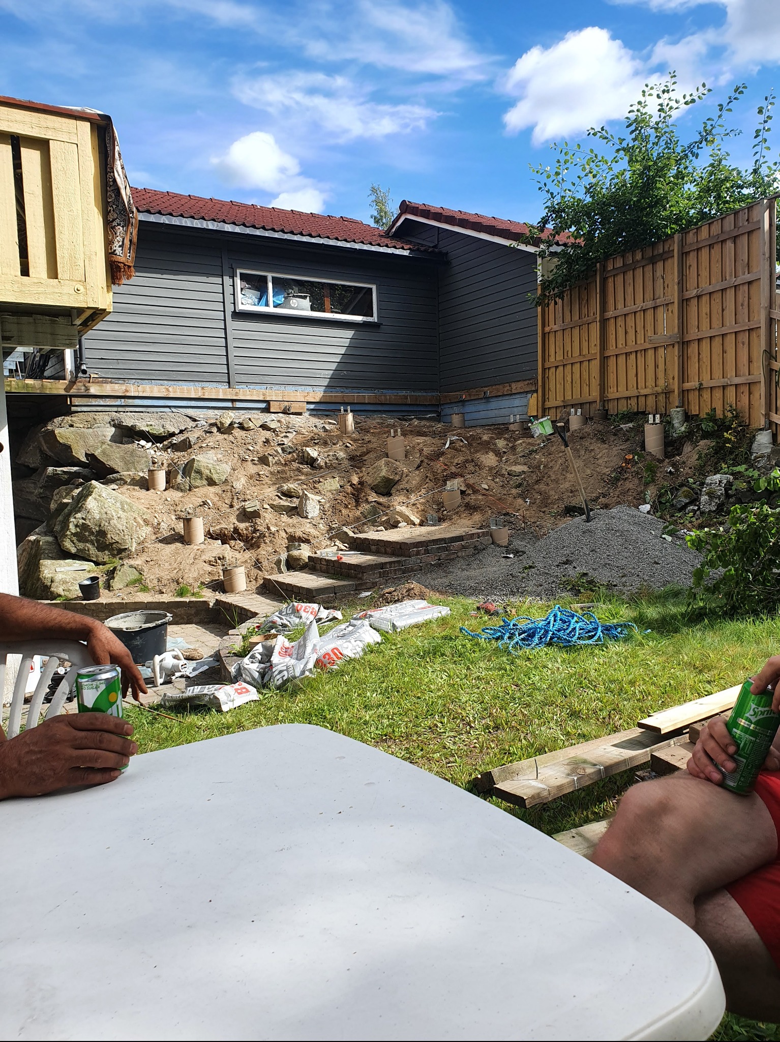 Przygotowania do budowy tarasu: fundamenty z betonowych słupów, ziemia, kamienie, narzędzia. Przerwa na napój, widoczne ręce z puszkami. Fragment domu z drewnianą elewacją.