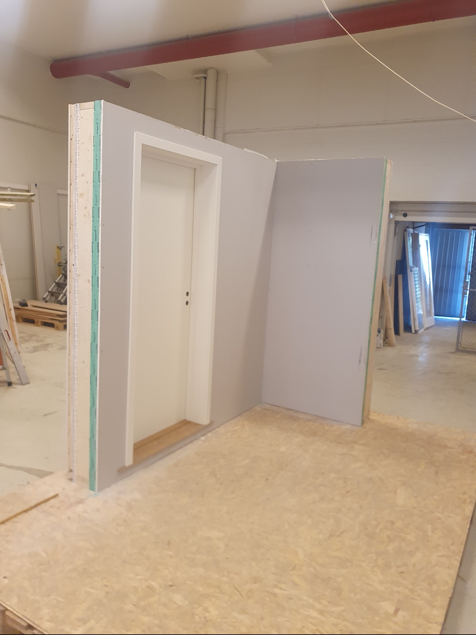 Wnętrze w trakcie prac: fragment ściany z drzwiami i płytą karton gips, podłoga z płyt OSB. Widoczne elementy konstrukcyjne i wykończeniowe. Prace wykończeniowe w toku.