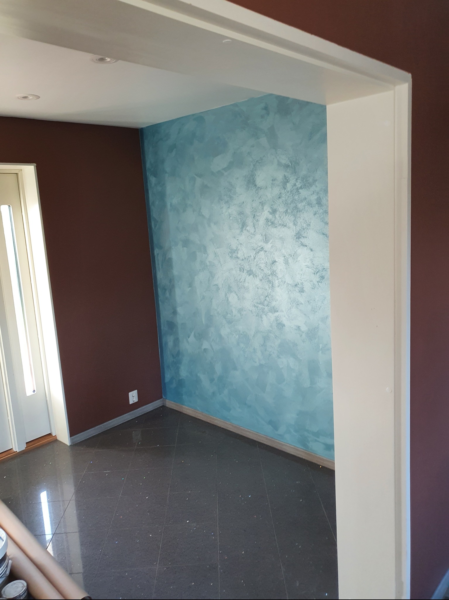 Pokój po malowaniu: ściana w odcieniach błękitu z efektem dekoracyjnym, brązowa ściana obok drzwi, ciemna podłoga z połyskiem. Widok z perspektywy wejścia.