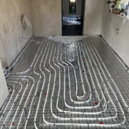 Białe rury ogrzewania podłogowego ułożone na czarnej siatce w surowym wnętrzu. Widok na sąsiednie pomieszczenie z analogiczną instalacją. Detale: czerwone znaczniki.