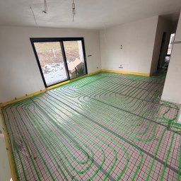 ARTUR BARAN INSTALACJE GRZEWCZE I SANITARNE - Zielone rury ogrzewania podłogowego ułożone na czerwonej siatce w surowym wnętrzu z oknem i żółtą taśmą brzegową. Widok z góry na instalację przed wylaniem posadzki.