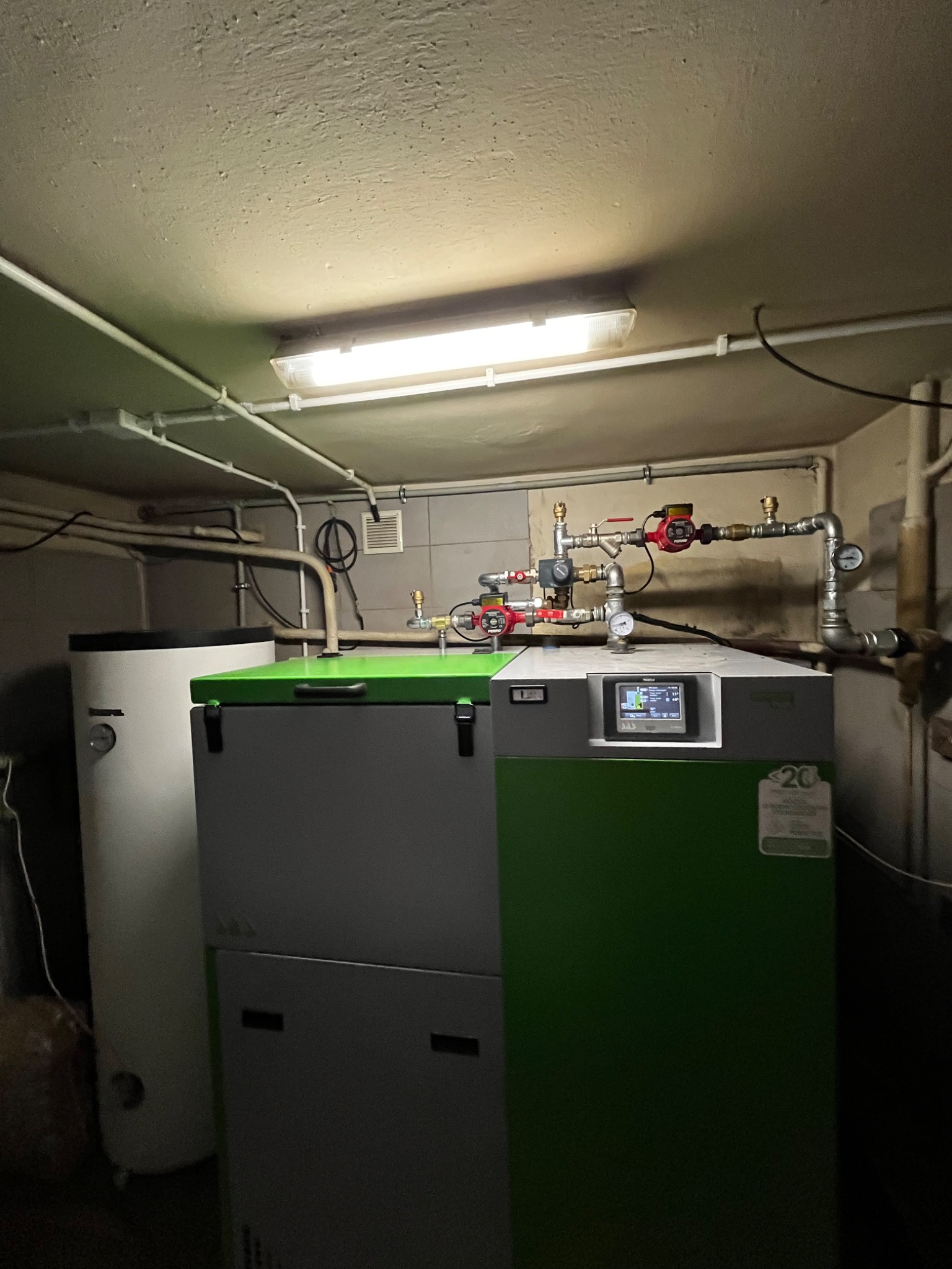 Nowoczesny piec grzewczy z zielonymi akcentami, podłączony do instalacji z pompami obiegowymi i zbiornikiem, oświetlony lampą jarzeniową w pomieszczeniu technicznym.