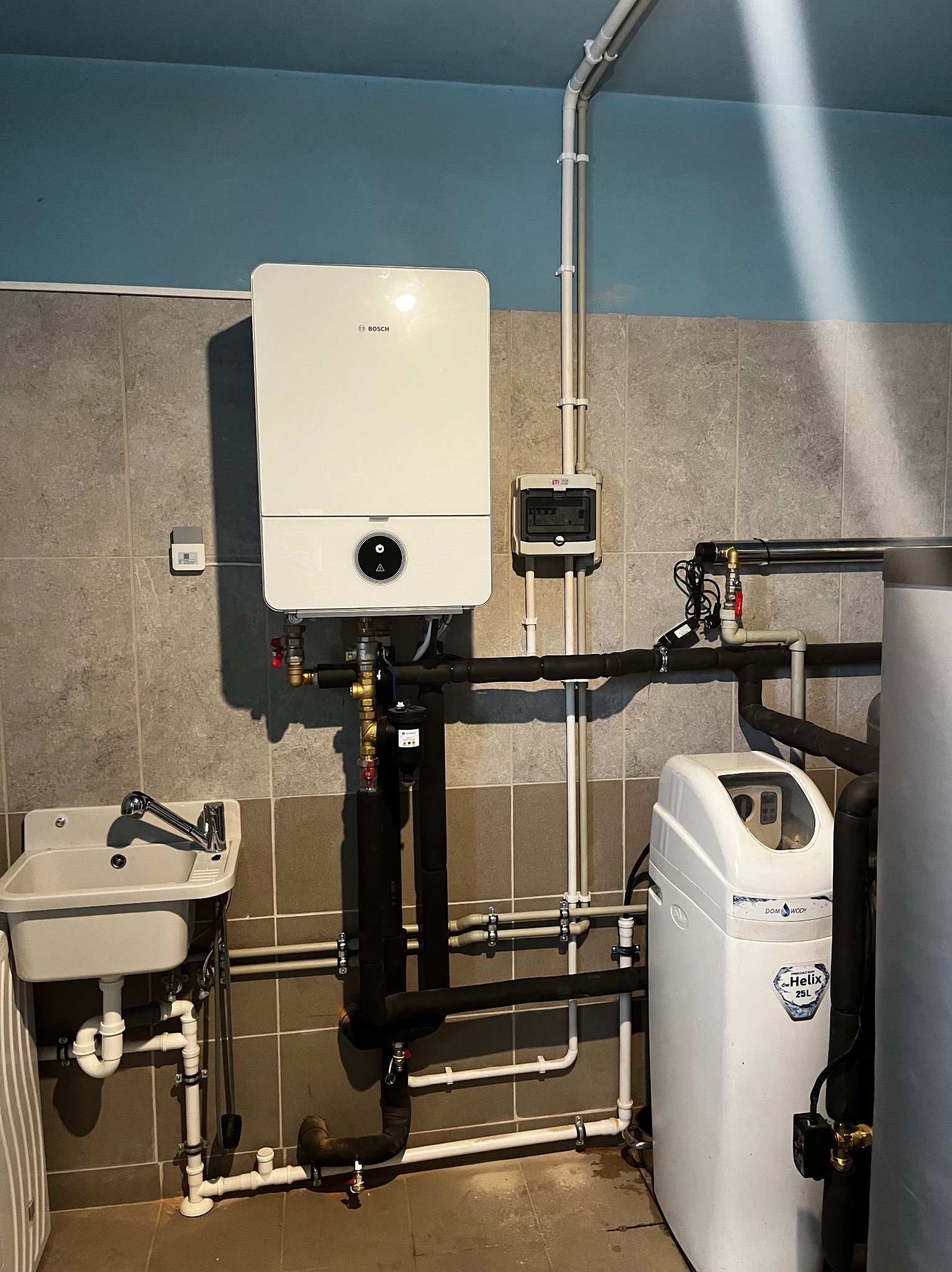 Nowoczesny piec gazowy Bosch, filtr do wody Dom Wody Helix 25L oraz umywalka w pomieszczeniu z szarymi płytkami i niebieską górną częścią ściany.