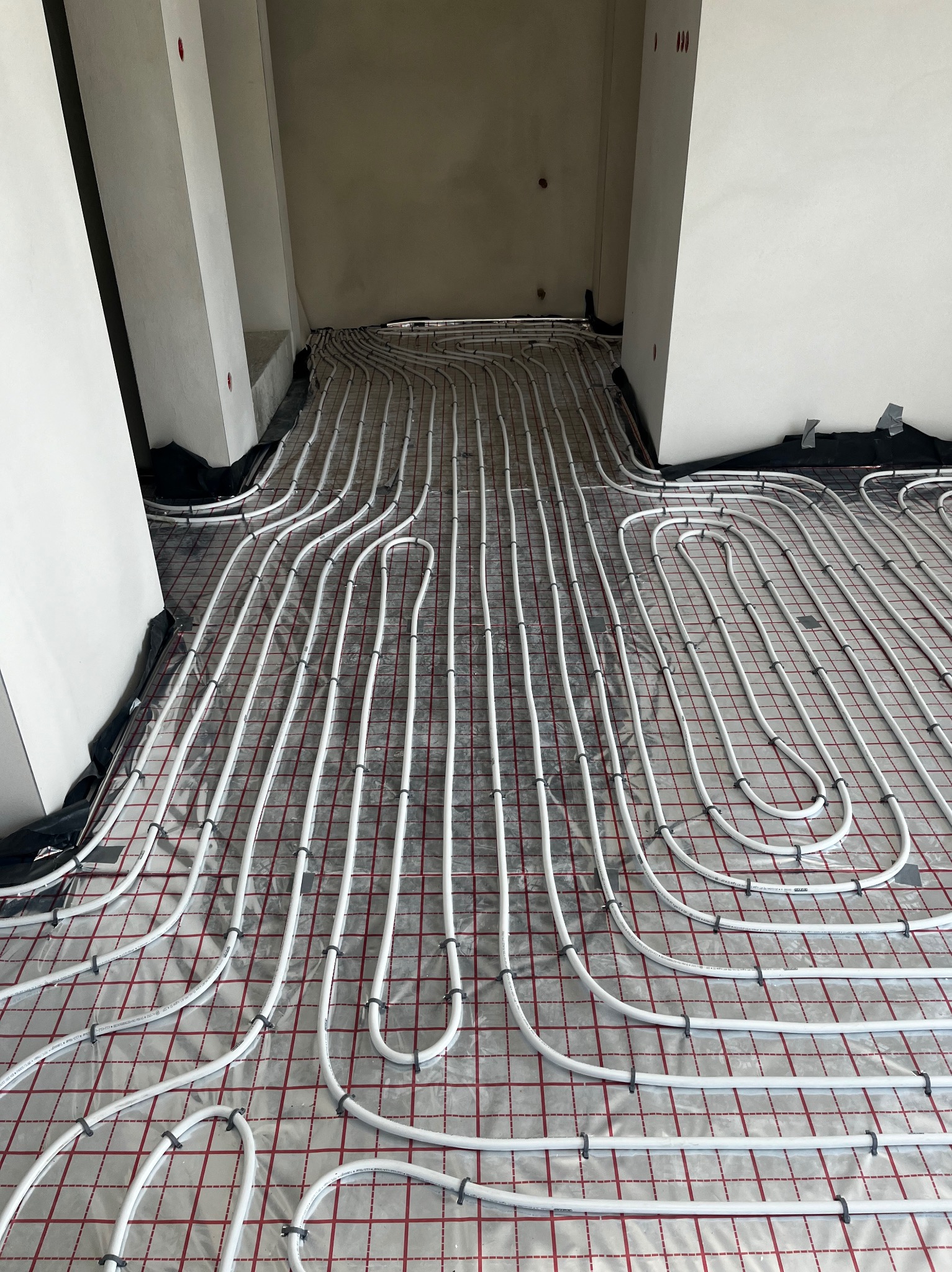 Białe rury ogrzewania podłogowego ułożone na folii z czerwoną kratką w trakcie montażu w Nowym Sączu, widoczne mocowania rur. Surowy stan ścian.