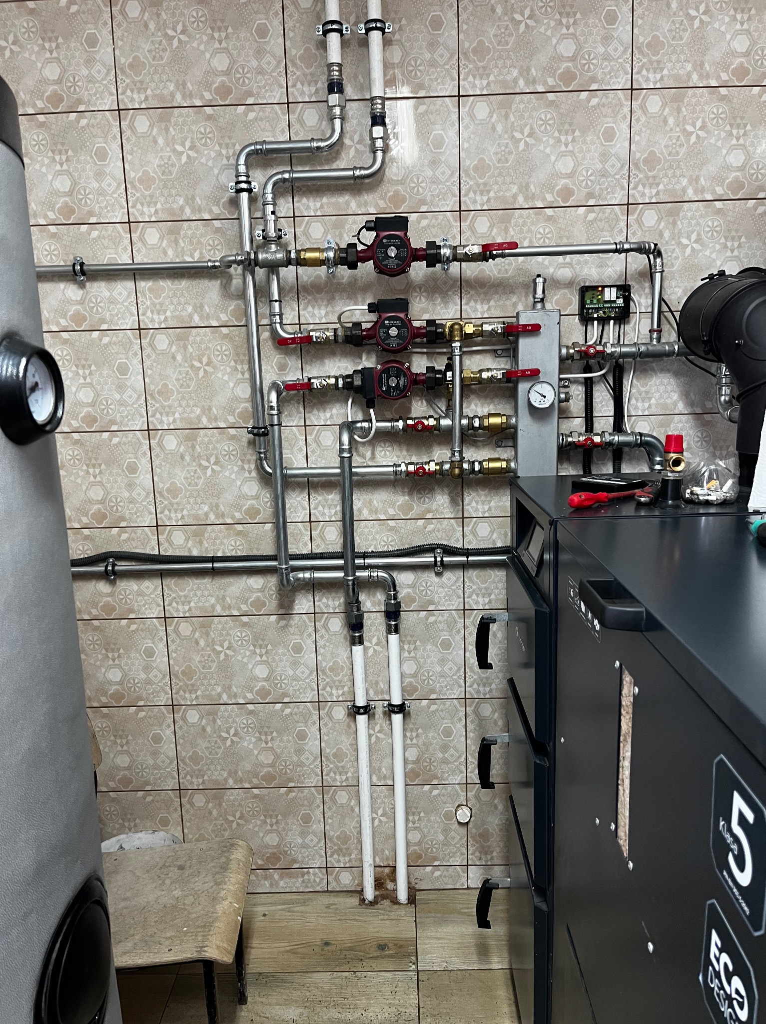 Złożona instalacja grzewcza z piecem na paliwo stałe, widoczne rury, pompy obiegowe, zawory i sterownik na tle płytek ceramicznych