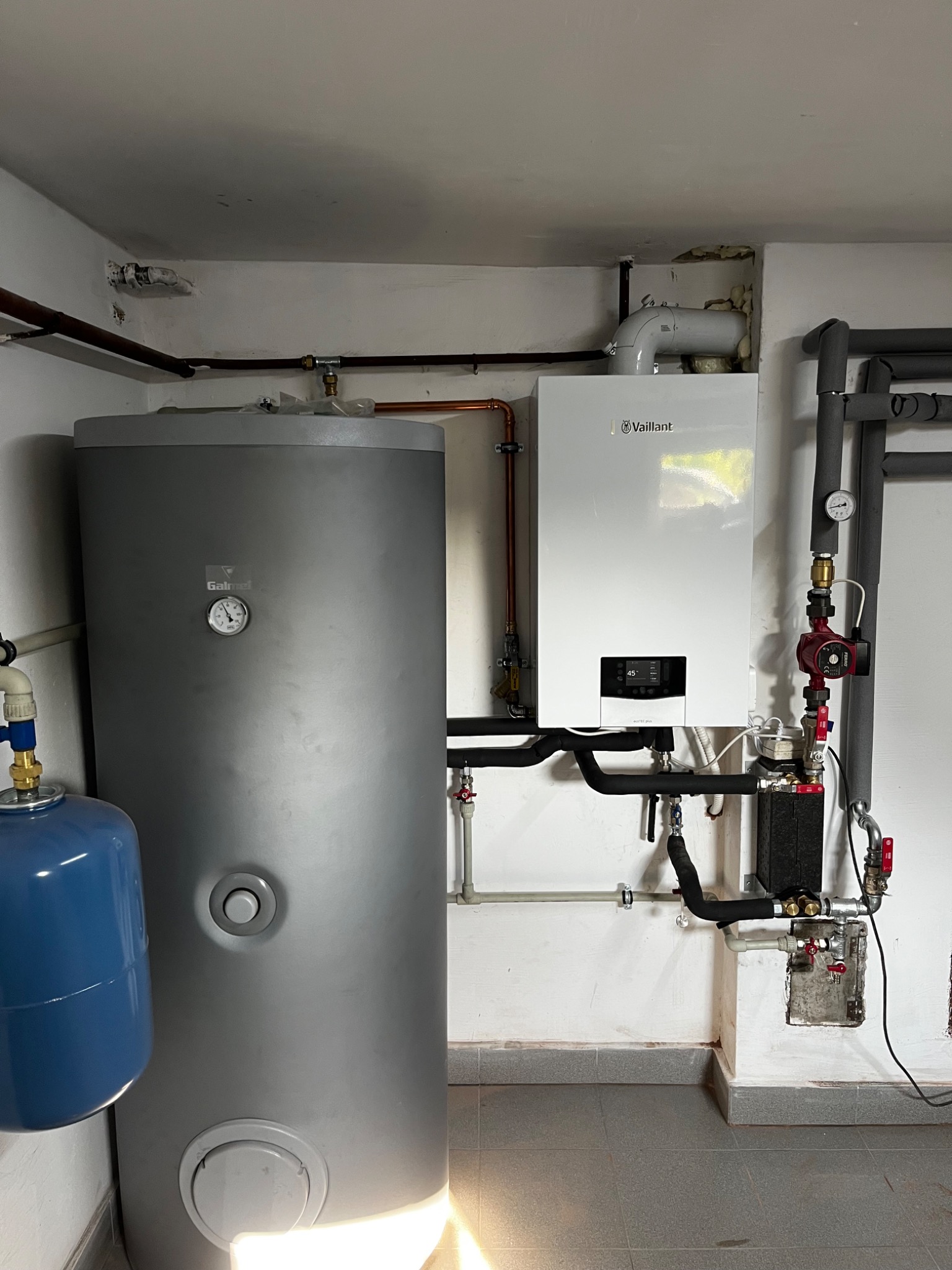 Nowoczesny piec gazowy Vaillant z zasobnikiem Galmet, zainstalowany w pomieszczeniu z szarą podłogą i białymi ścianami, po prawej widoczne rury instalacji grzewczej.