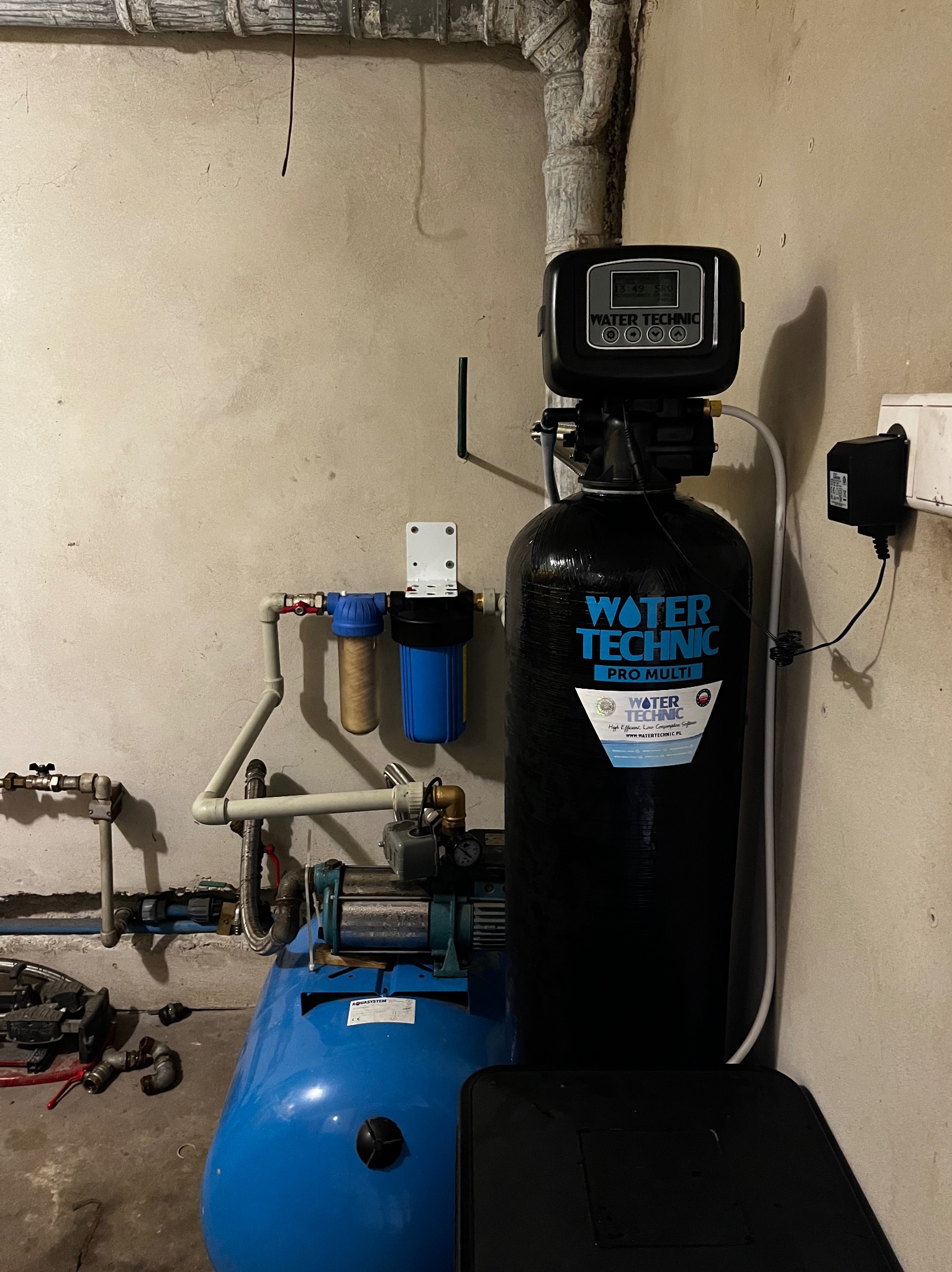 Zestaw hydroforowy z filtrami i zmiękczaczem wody Water Technic Pro Multi w piwnicy. Widoczne rury, pompy i elementy instalacji w szarej piwnicy.