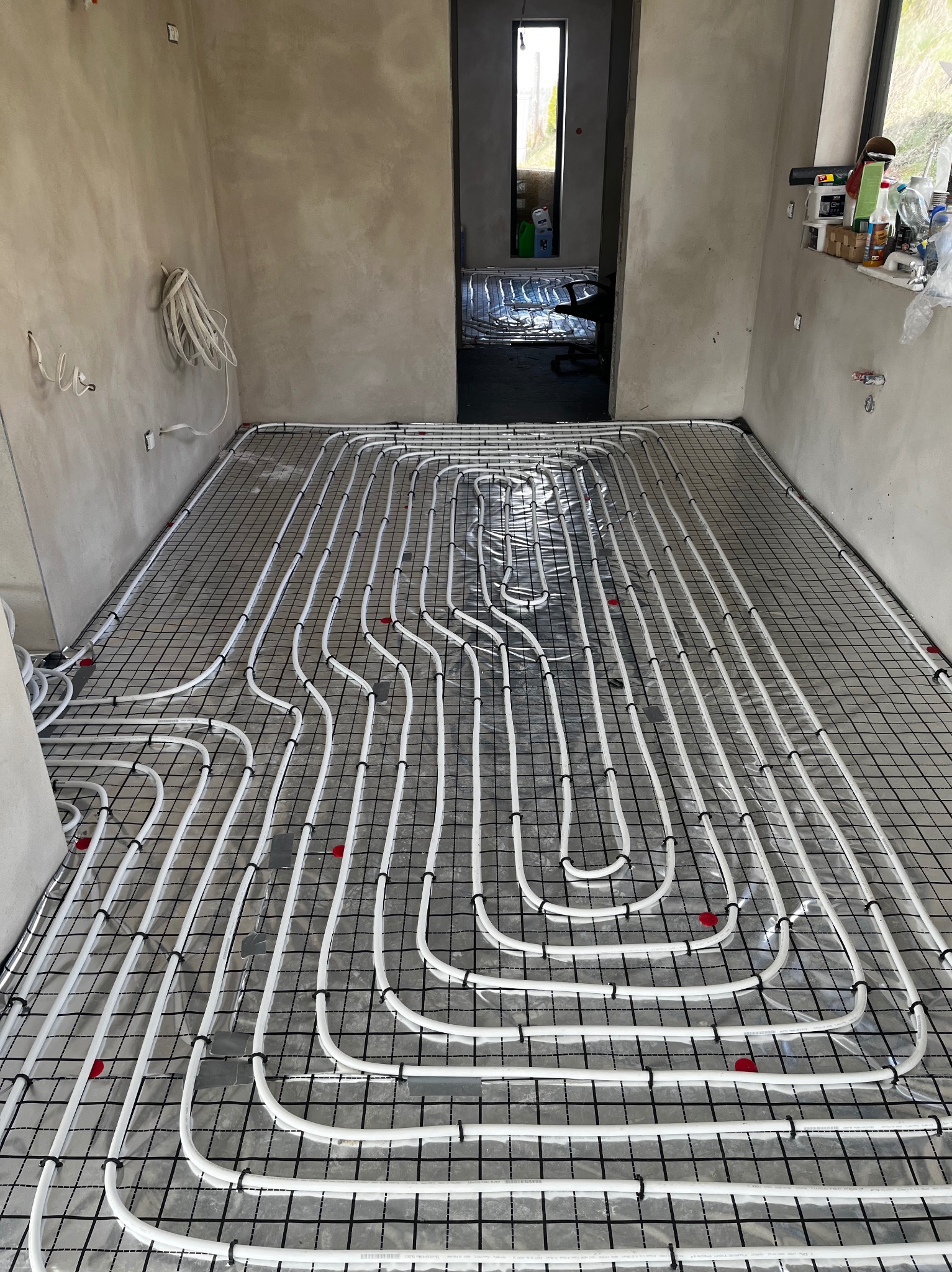 Białe rury ogrzewania podłogowego ułożone na czarnej siatce w surowym wnętrzu. Widok na sąsiednie pomieszczenie z analogiczną instalacją. Detale: czerwone znaczniki.