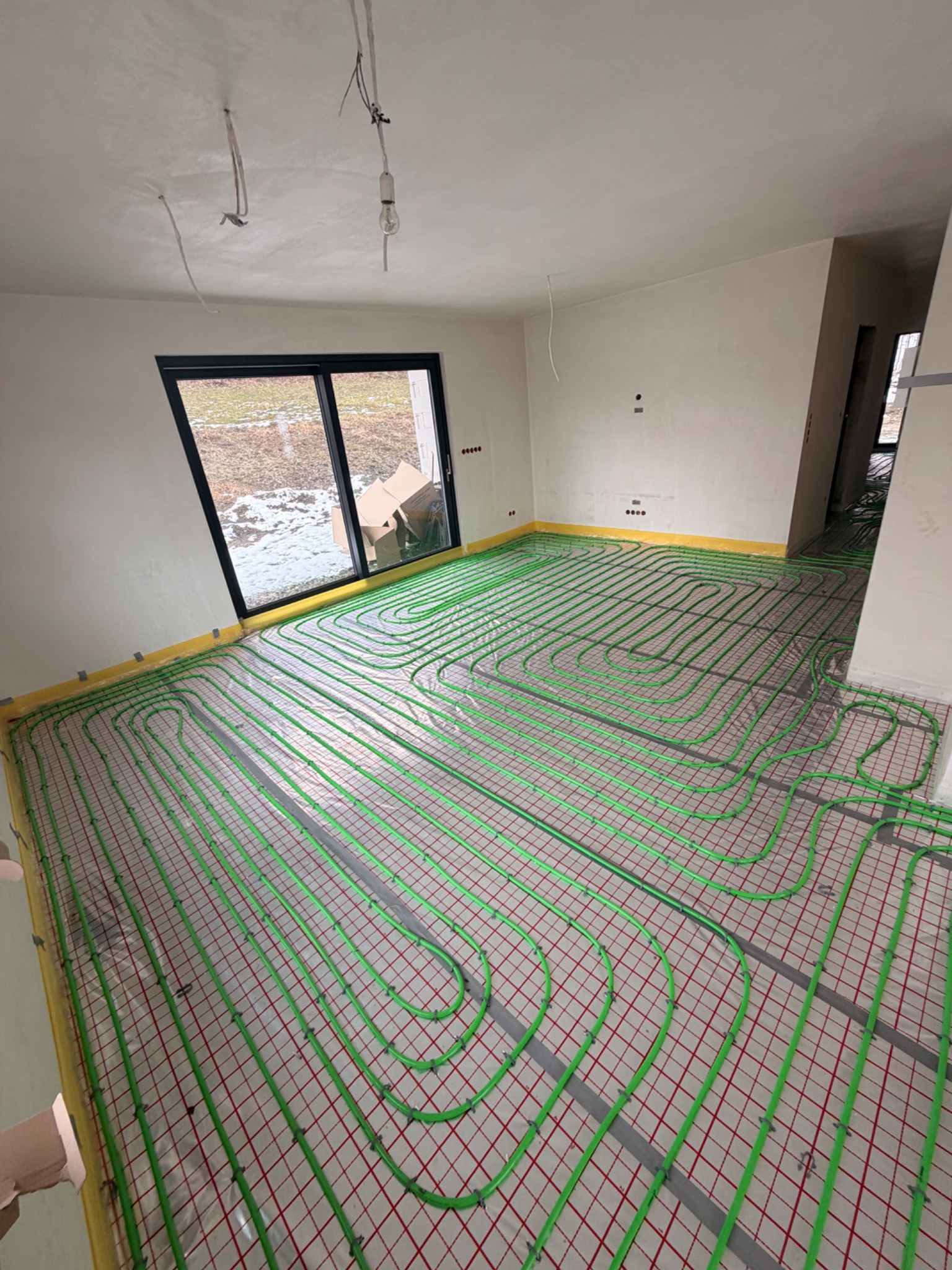 Zielone rury ogrzewania podłogowego ułożone na czerwonej siatce w surowym wnętrzu z oknem i żółtą taśmą brzegową. Widok z góry na instalację przed wylaniem posadzki.