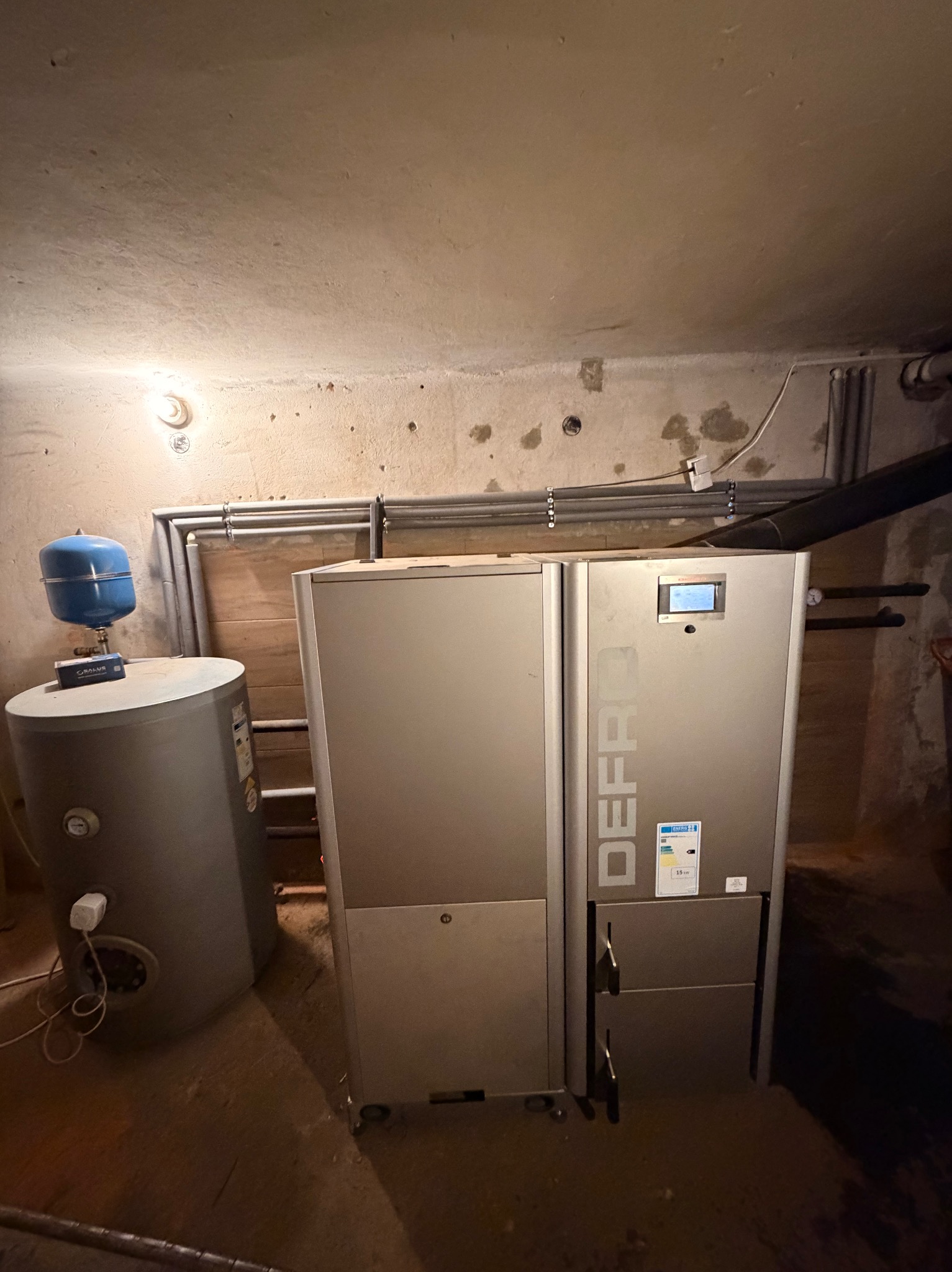 Szary piec CO marki Defro z podłączonym zbiornikiem wyrównawczym w kotłowni. Widoczne rury instalacji grzewczej i oświetlenie. Niska jakość zdjęcia.