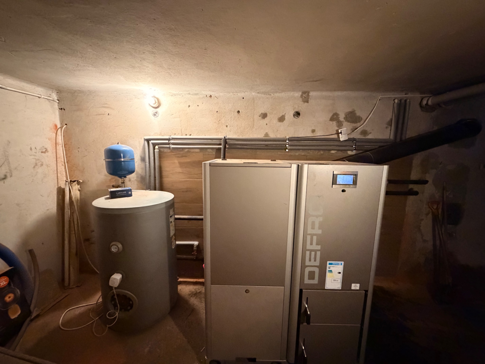 Nowoczesny piec CO marki Defro w kotłowni z zbiornikiem wyrównawczym i instalacją. Widoczne rury i surowy beton. Oświetlenie górne.