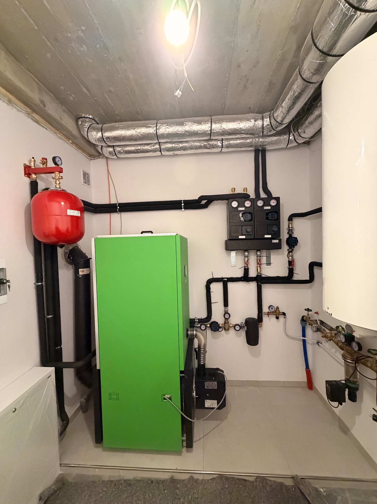 Nowoczesna kotłownia z zielonym piecem w Nowym Sączu. Widoczne rury, zbiornik wyrównawczy i osprzęt. Instalacja grzewcza w pomieszczeniu z surowym betonowym stropem.