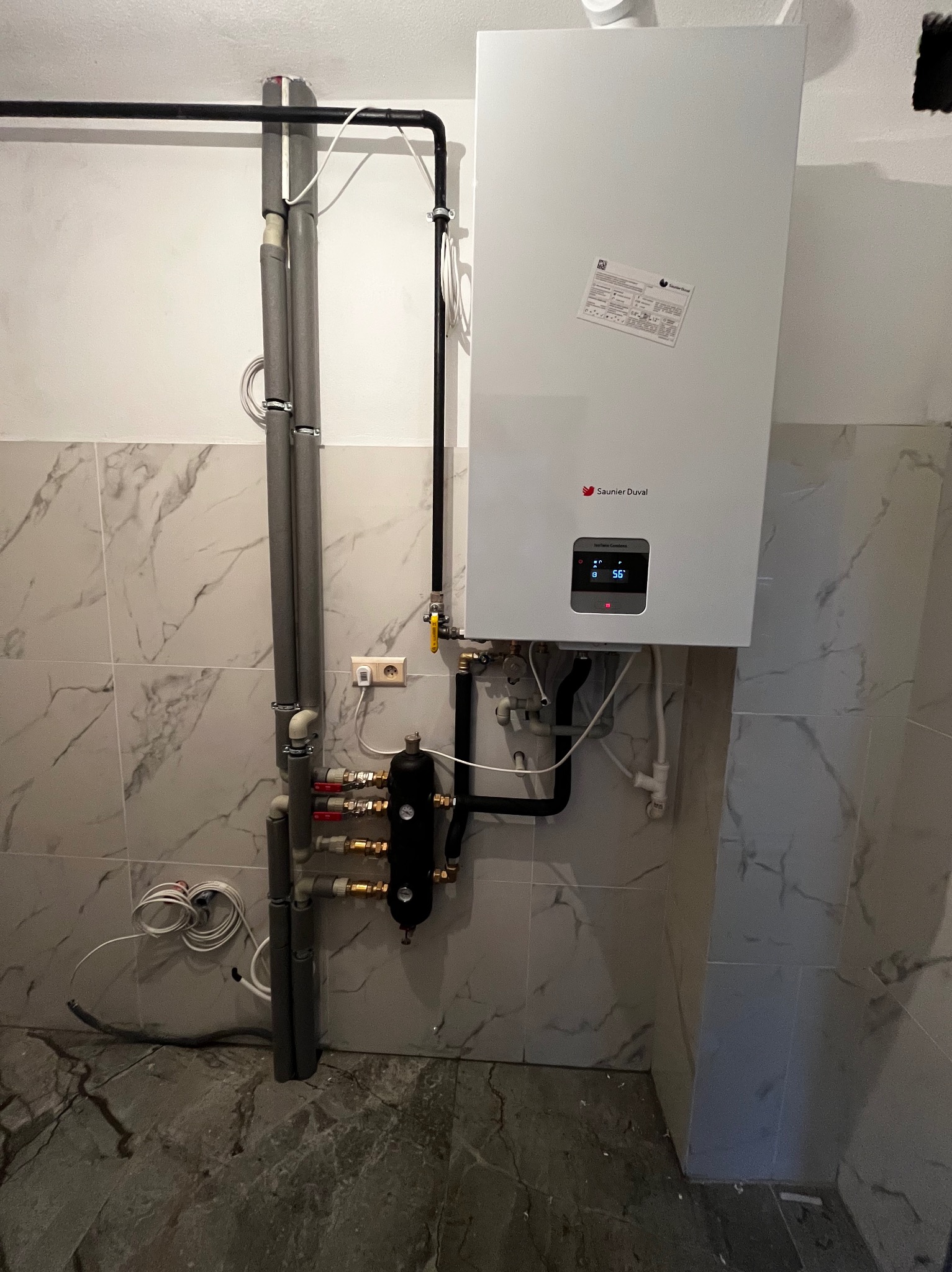Biały piec gazowy Saunier Duval zamontowany na ścianie z marmurowymi płytkami. Widoczne rury, zawory i czarny filtr. Instalacja w pomieszczeniu z szarą podłogą.